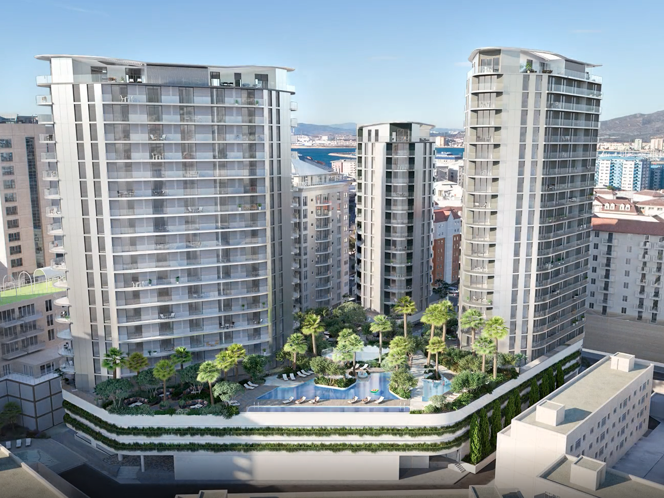 Aerial view of modern high-rise residential buildings with a rooftop pool and garden area, situated in an urban city with mountains and water in the background.