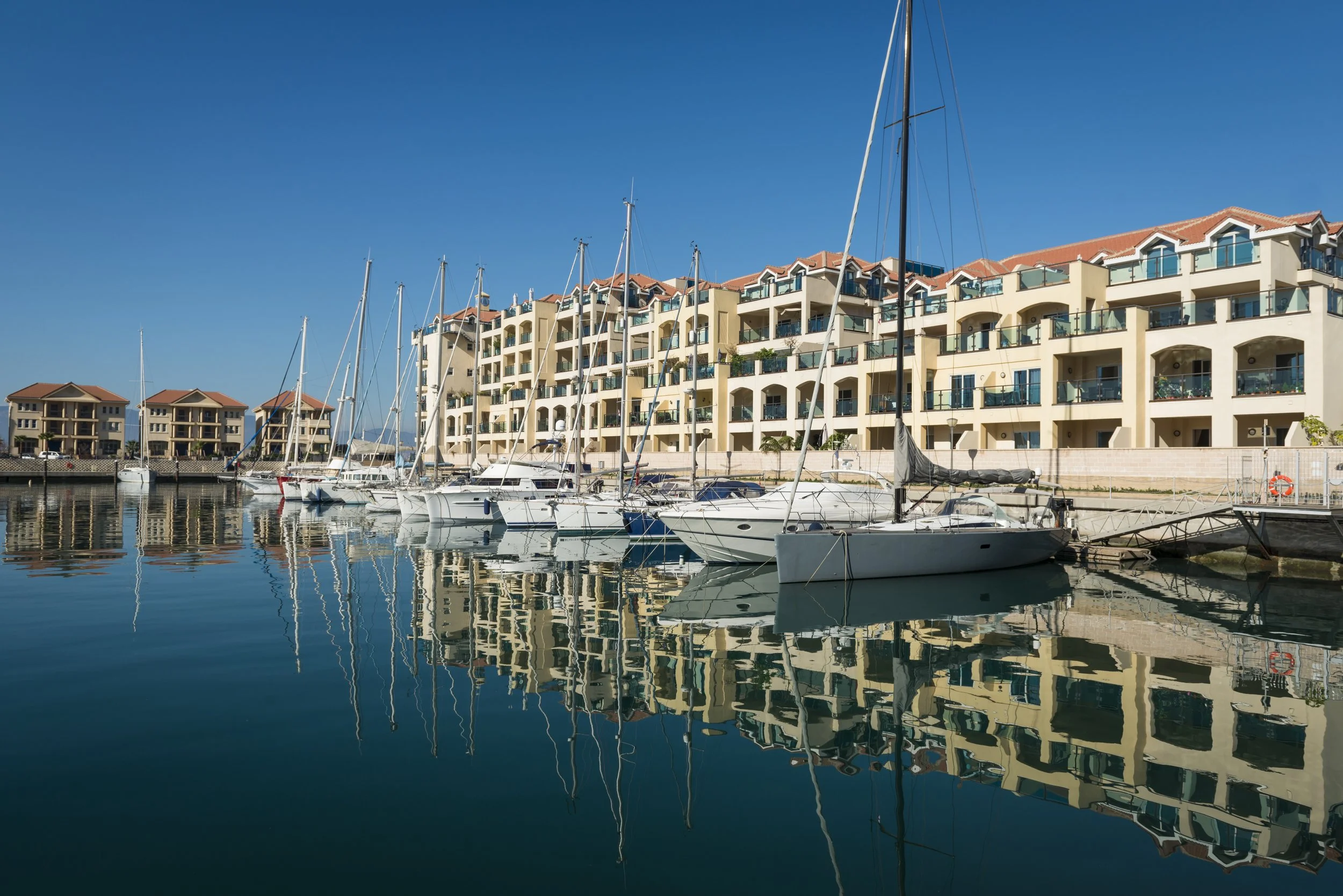 A marina with several sailboats moored in calm water, and modern beige apartment buildings with red-tiled roofs in the background under a clear blue sky.