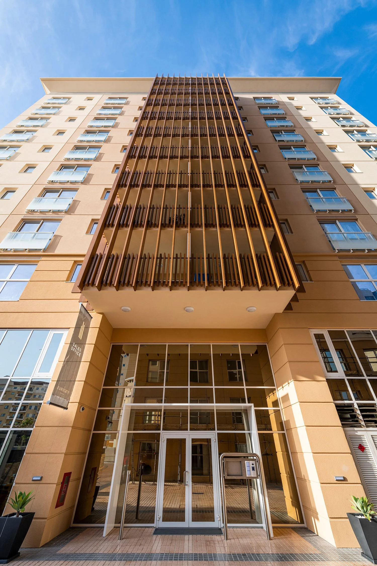 Apartment building with glass doors at entrance, metal turnstile, and potted plants, under a blue sky.