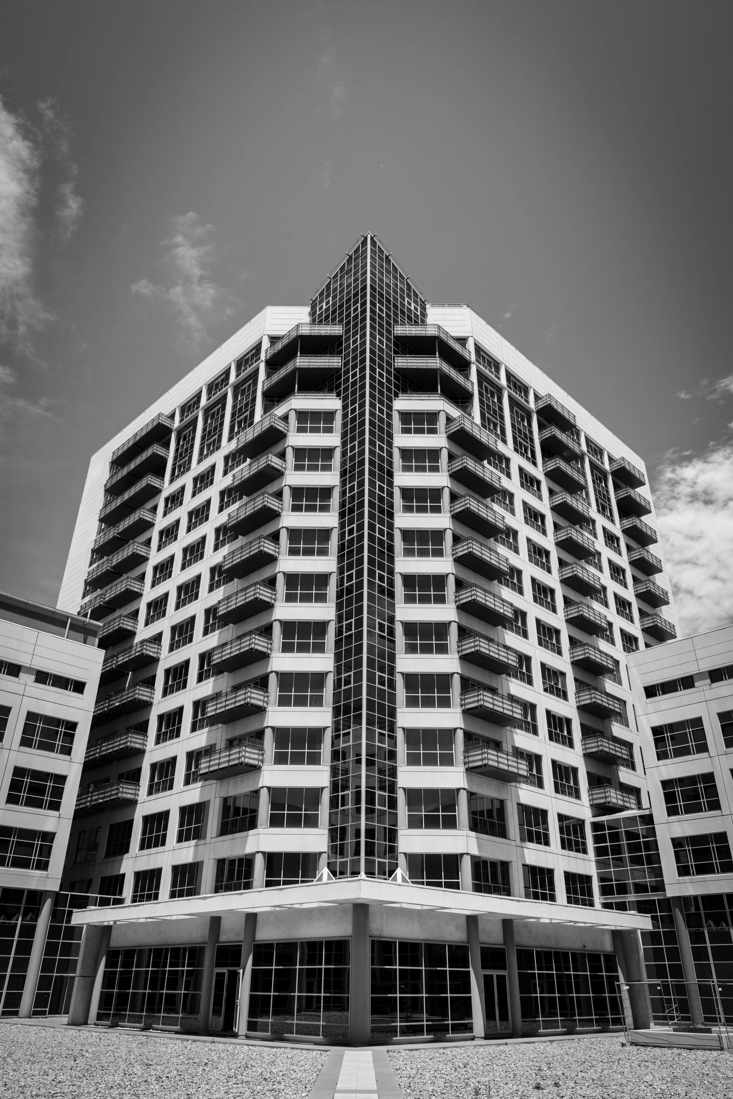 A modern multi-story building with many balconies and large glass windows, viewed from ground level looking up, with a cloudy sky in the background.