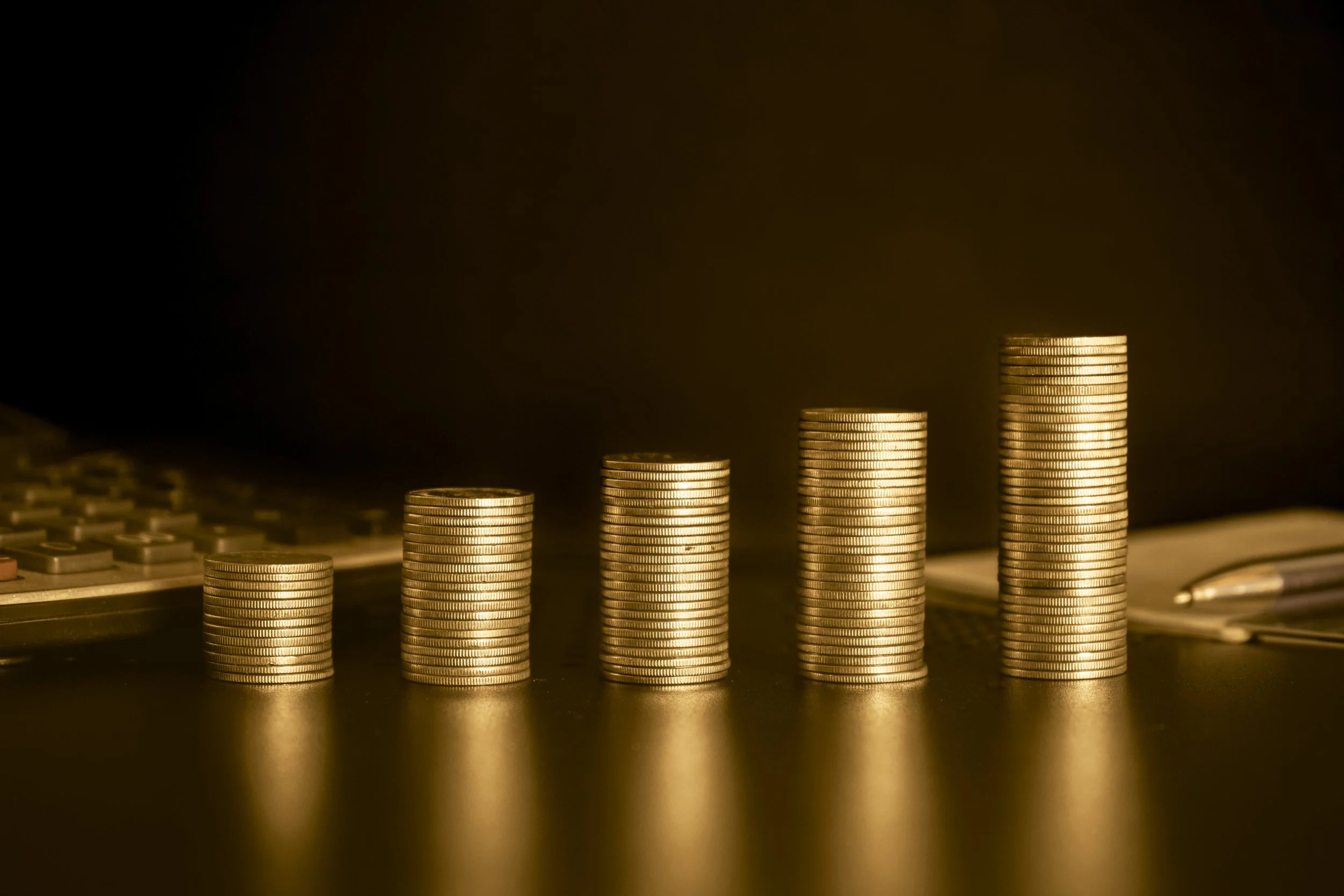Stacks of coins arranged in increasing height on a dark surface with a calculator in the background.