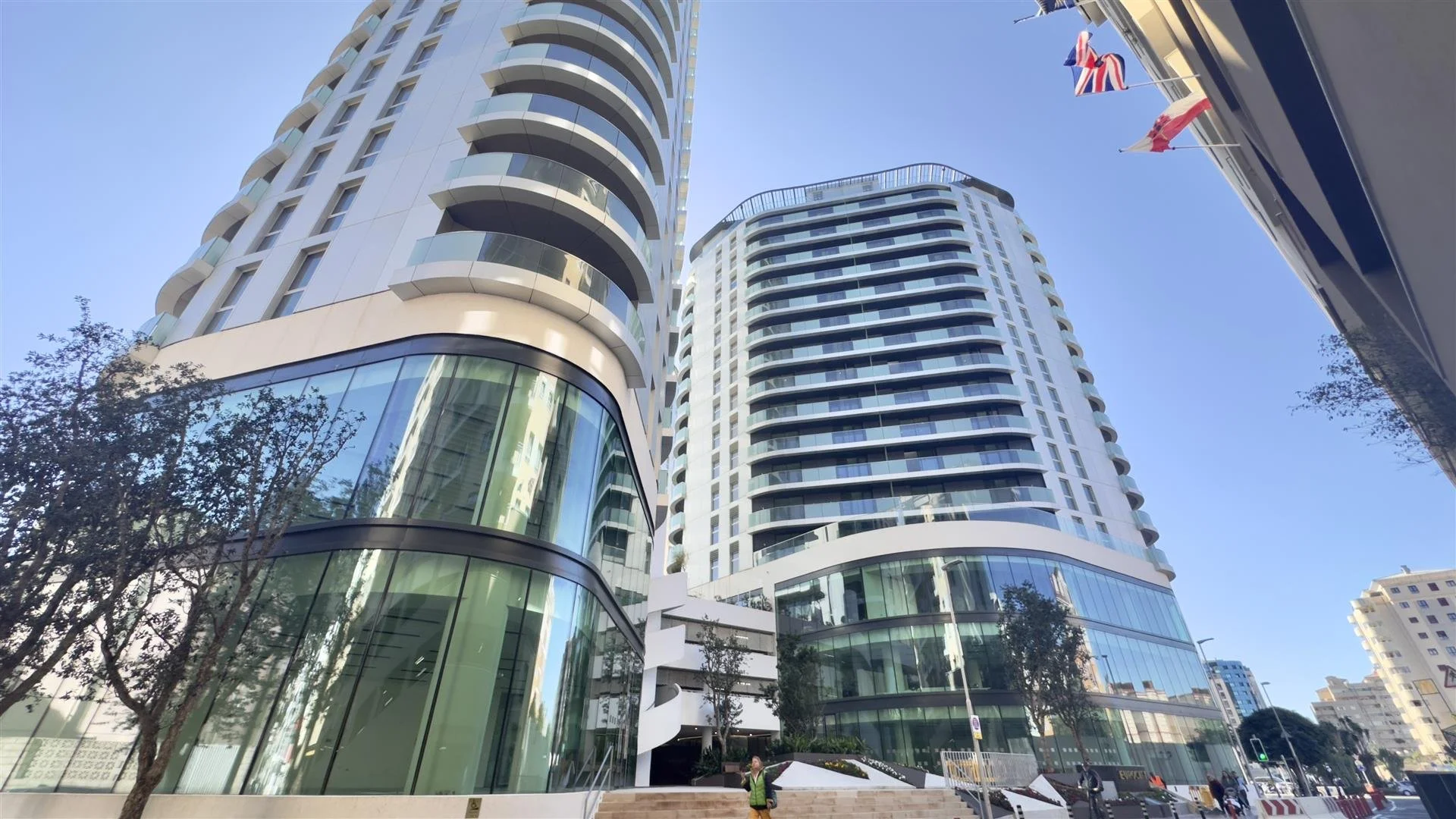 Low-angle view of modern high-rise buildings with glass facades and curved balconies, set against a clear blue sky. Flags are visible on the right side of the image, and a person in a yellow vest is at the base, near stairs and trees.