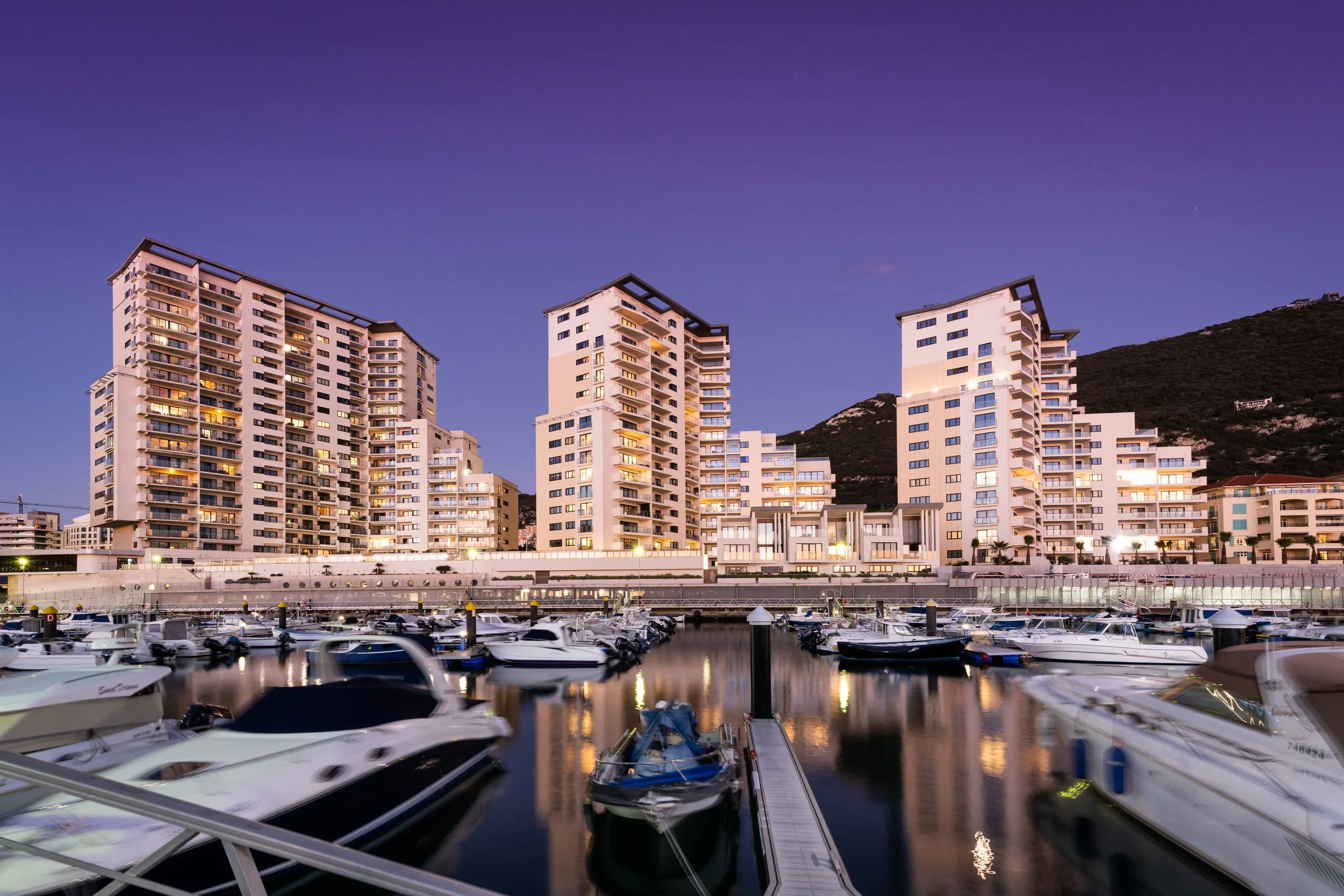Nighttime view of tall modern white residential buildings with lit windows, situated behind a marina filled with various boats docked at the docks, with a hillside in the background.