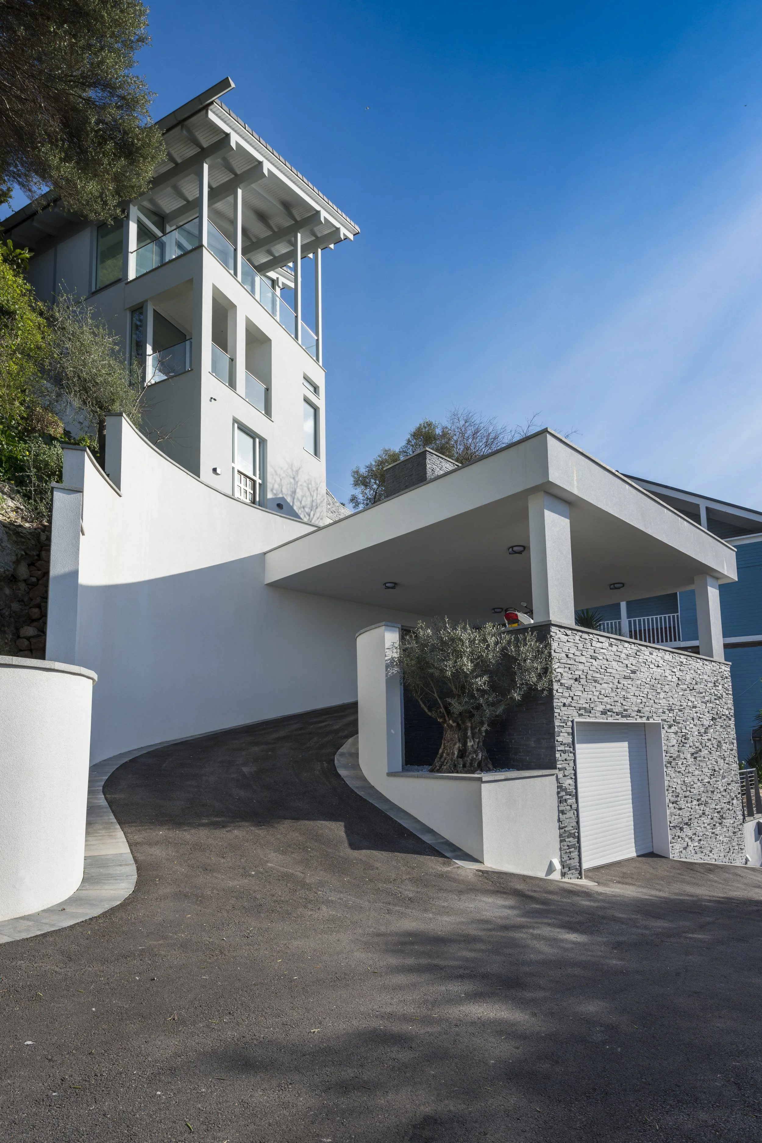 Modern multi-story house with white exterior walls, large windows, a driveway winding up a hill, and a garage door. There is a tree with twisted branches near the house and a clear blue sky overhead.