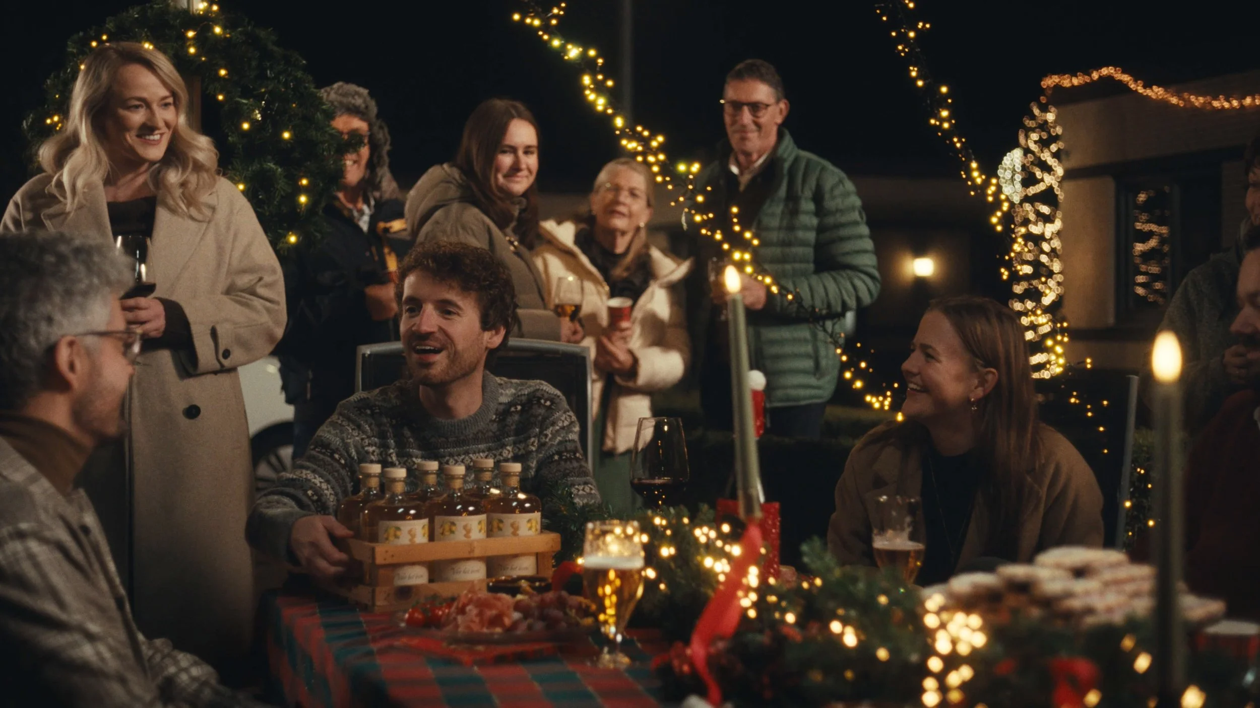 Een groep mensen viert feest tijdens een kerstfeest buiten 's avonds, met kerstverlichting en versieringen op de achtergrond. Ze lachen, hebben drankjes en eten, en creëren een feestelijke sfeer.