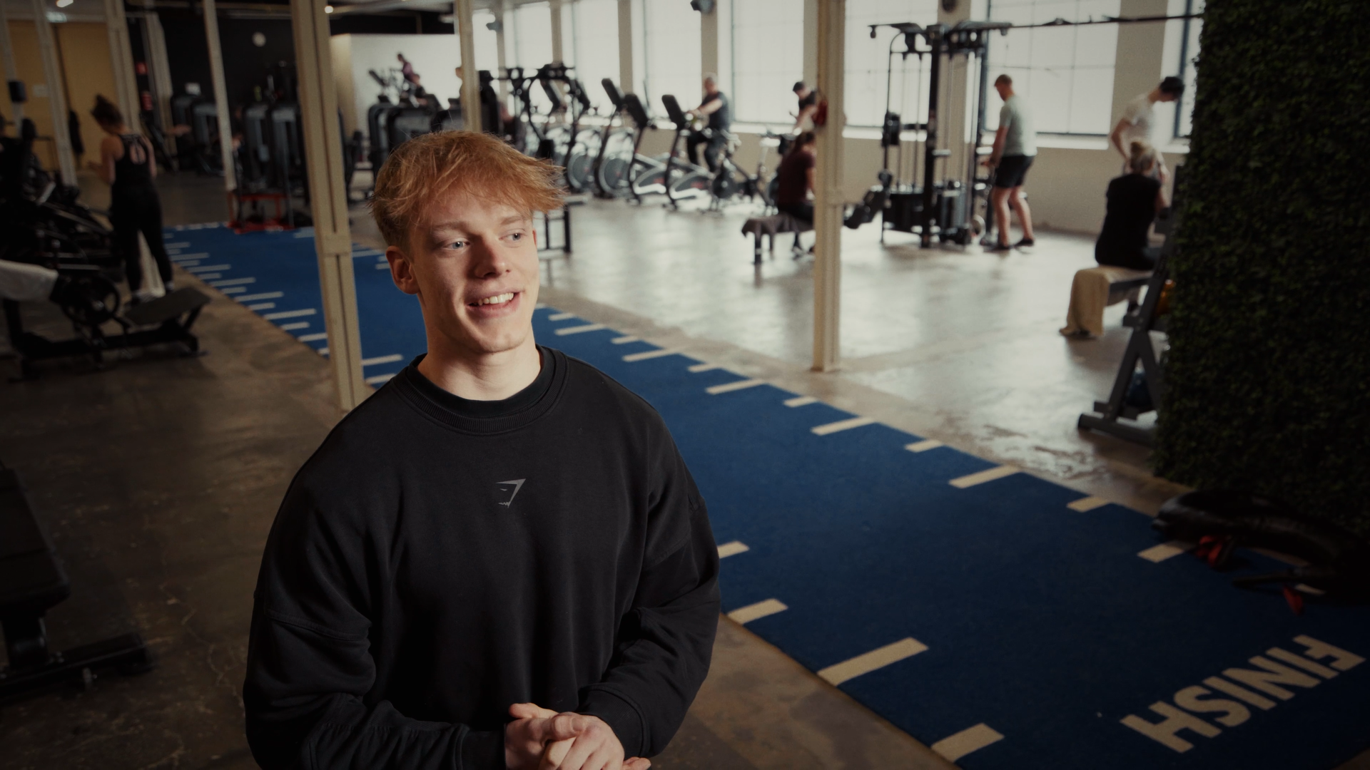 Een jonge man met rood haar in een sportschool, gekleed in een zwart sweatshirt, glimlachend en een gesprek op camera.
