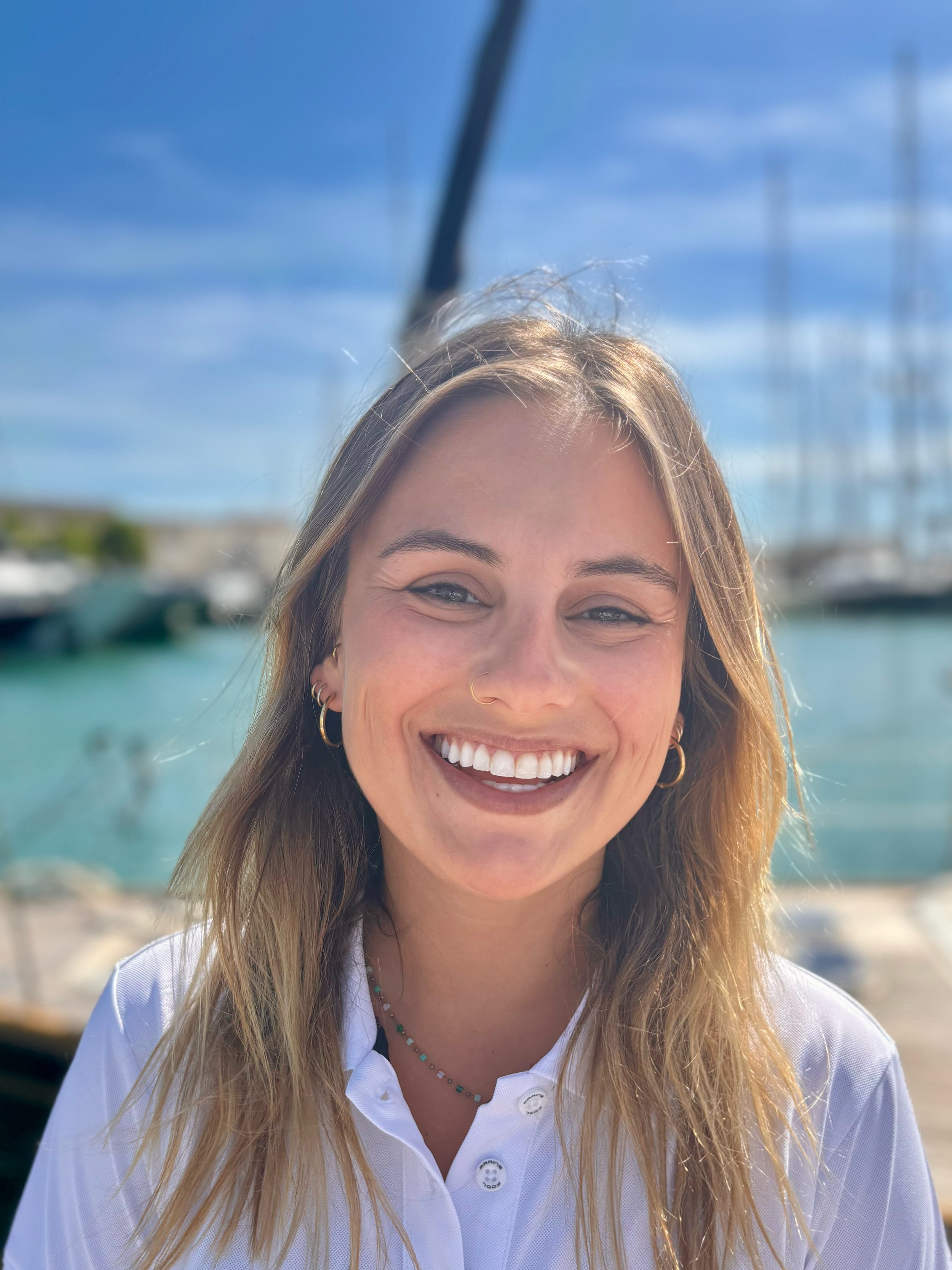 A smiling woman with long blonde hair, wearing a white shirt and jewelry, standing outdoors by a body of water with boats, under a blue sky.