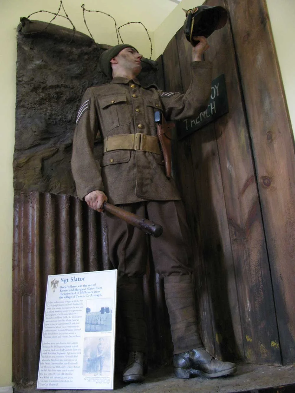 Life-sized statue of a WWII soldier in uniform, holding a helmet in one hand and a rifle in the other, placed in a museum setting.