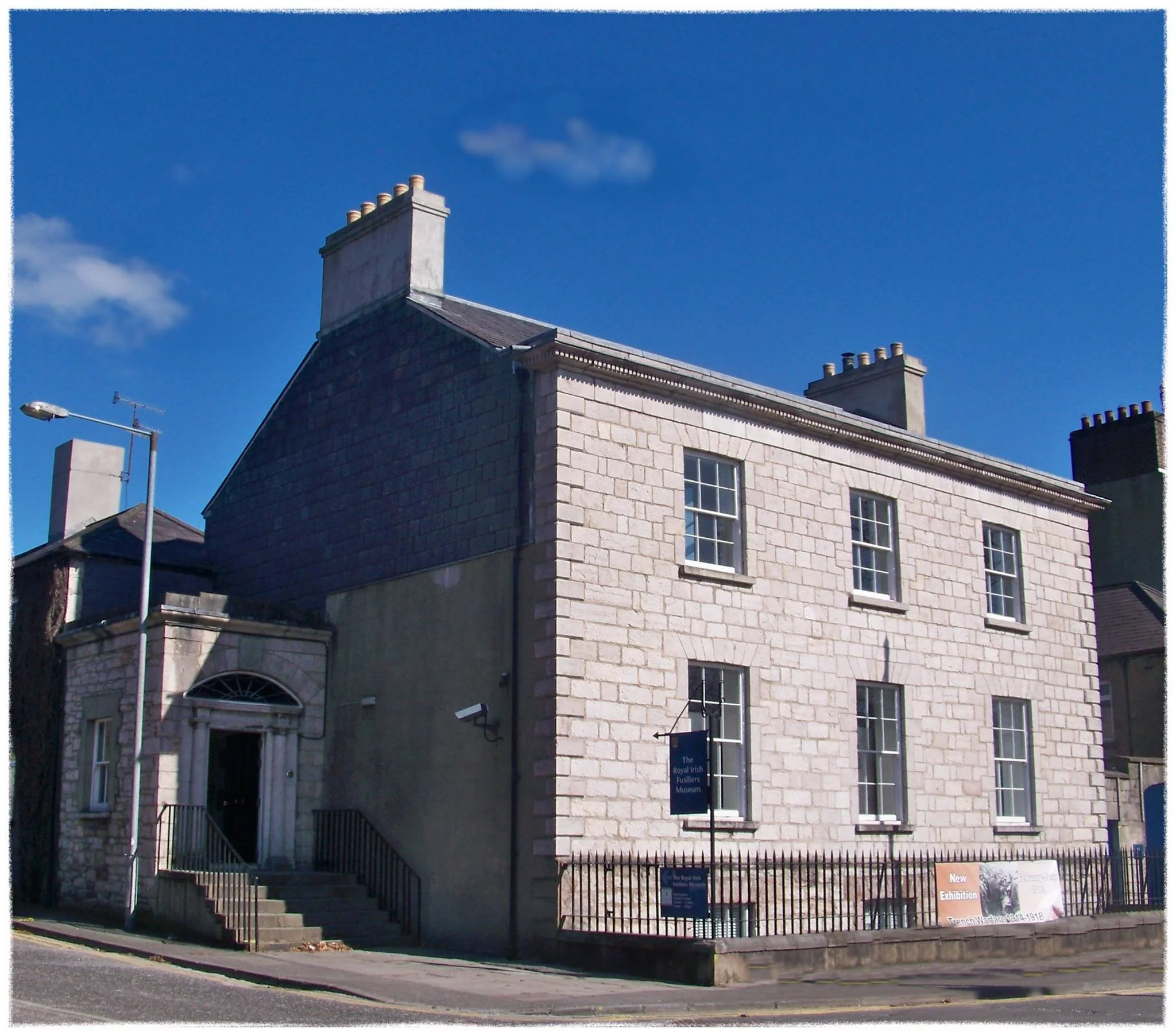 A historic stone building with a black shingled roof, multiple chimneys, and white-trimmed windows, situated against a bright blue sky.