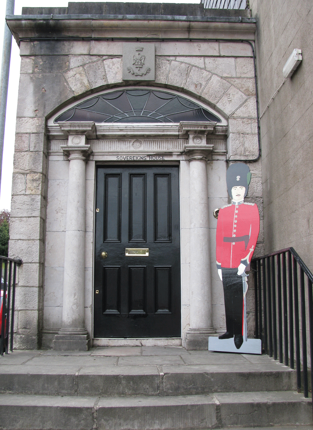 Entrance to Sovereign's House with a black door, stone columns, and a decorative arch above. A cutout statue of a British soldier dressed as a guard stands to the right of the door.