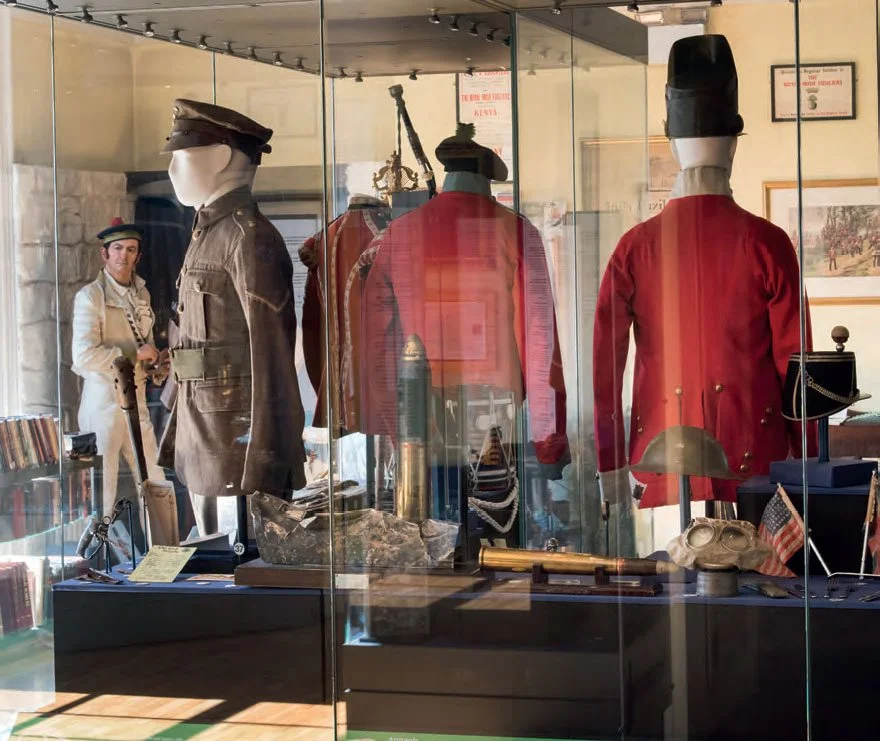 Display case with Civil War uniforms and artifacts, including a soldier's uniform, a hat, a sword, and a flag, with a person in period clothing in the background.
