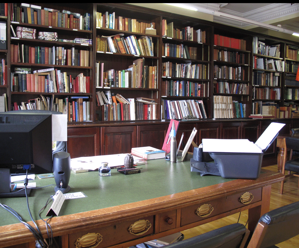An office with a green desk, a computer, a printer, and various office supplies, surrounded by tall wooden bookshelves filled with books.