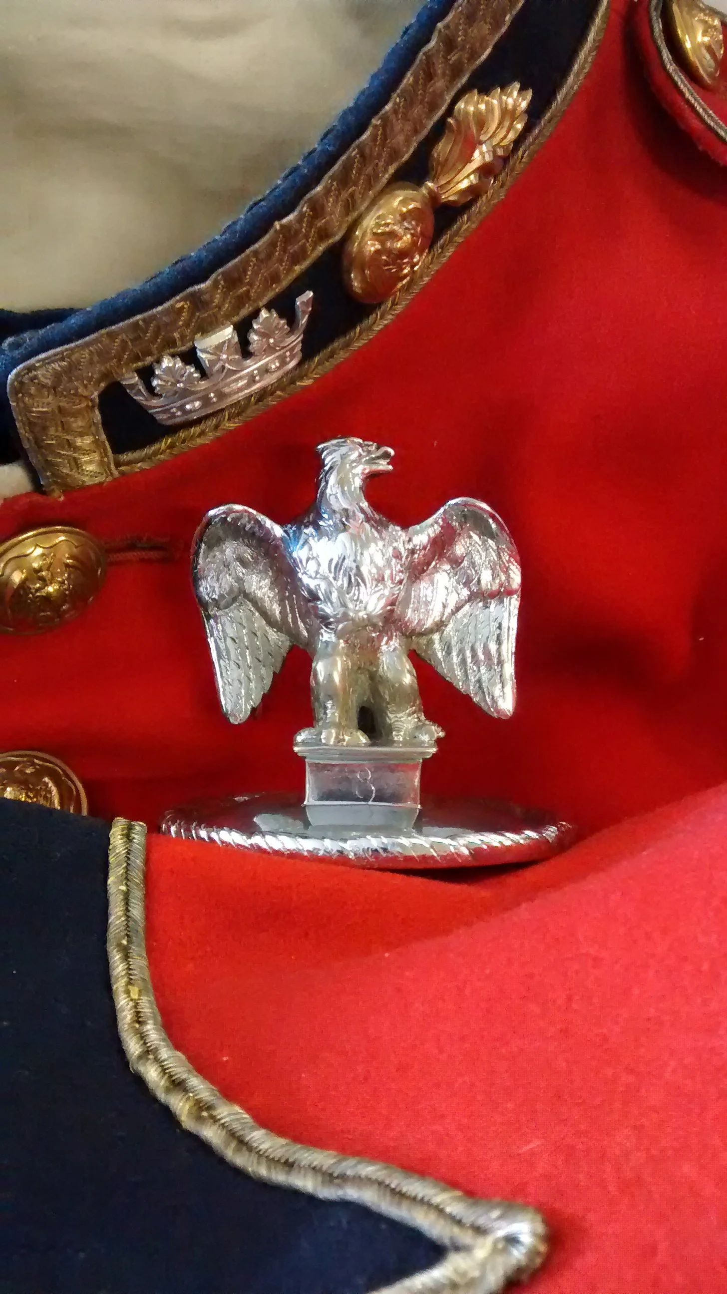 Close-up view of a silver eagle insignia on a red military uniform with decorative gold and black trim.