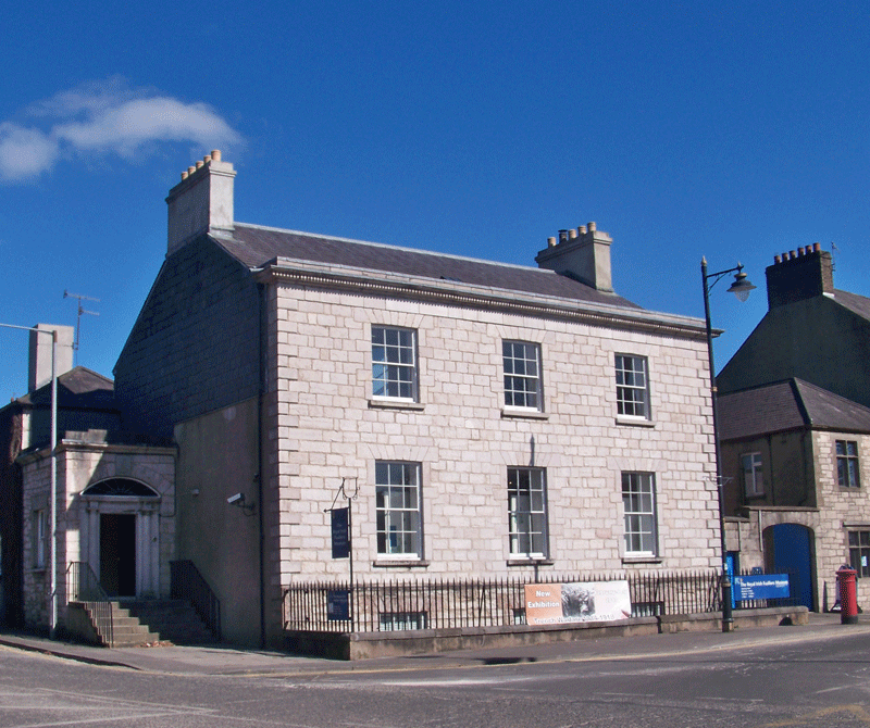 Stone building with multiple windows, small entrance with stairs, and a sign on the front, against a blue sky.
