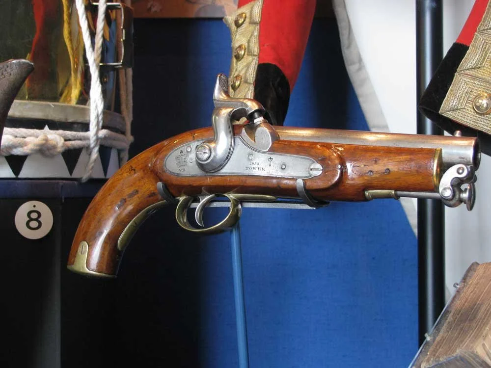 A historical cap and flintlock pistol with a wooden stock and brass fittings, displayed in a museum setting with historical clothing and props in the background.