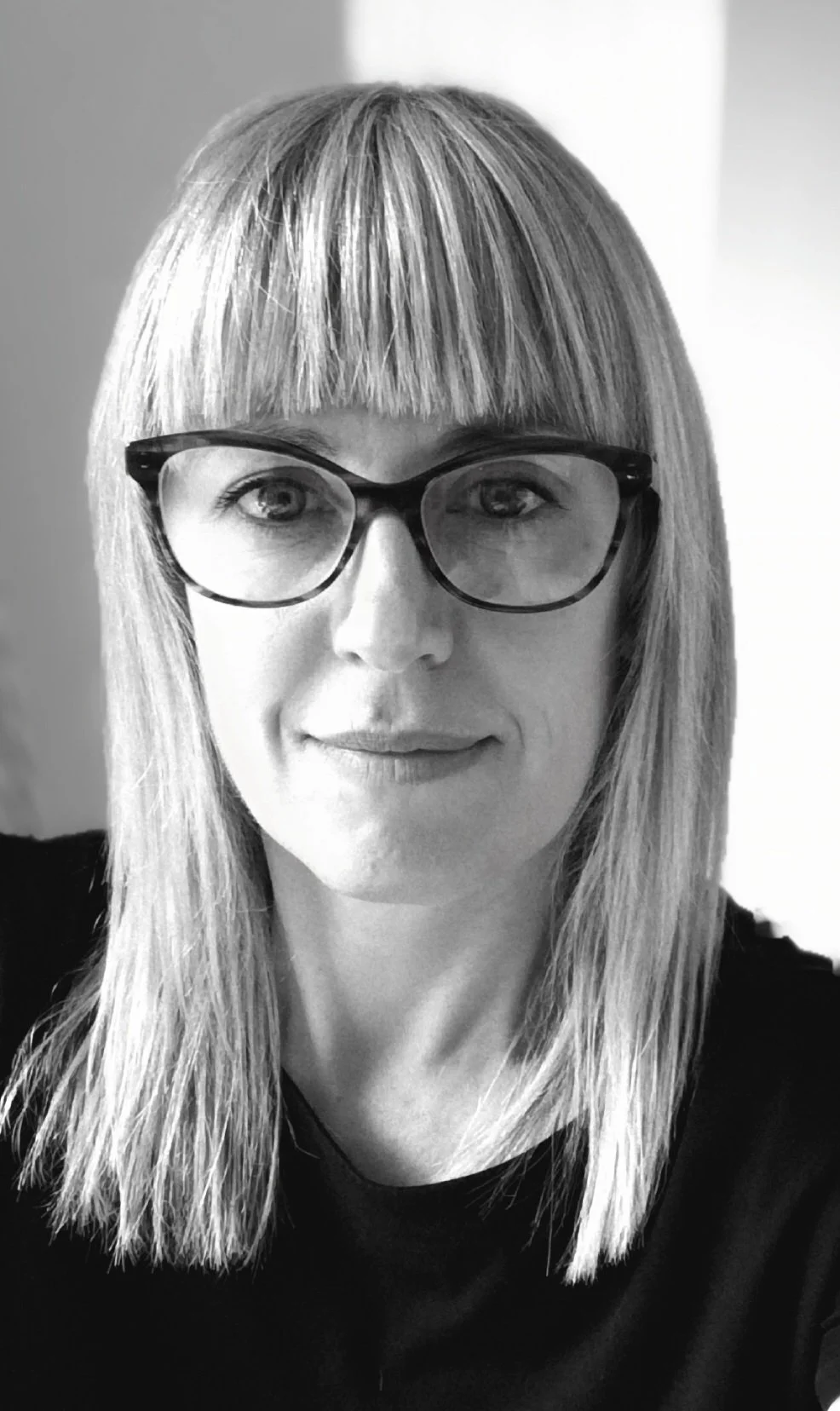 Black and white photograph of a woman with straight blonde hair, wearing large glasses and a dark top, smiling slightly.