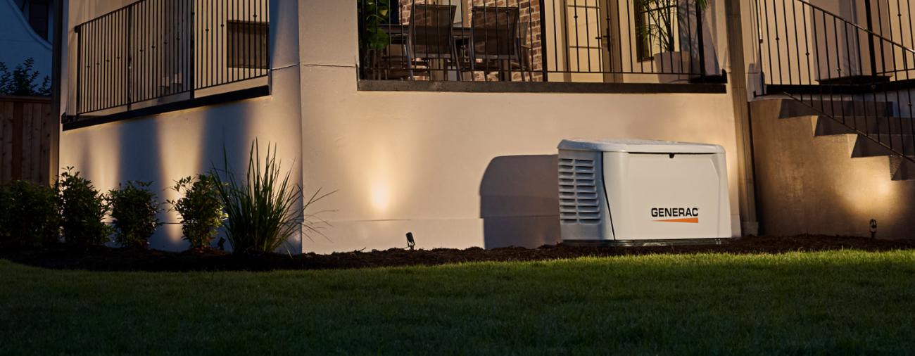 Outside view of a residential house at night with outdoor lighting illuminating the wall and stairs, a small landscaped garden with bushes and grass, and a Generac generator on the ground.