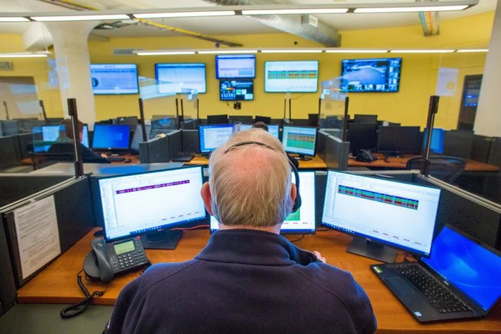 A man with gray hair and a dark jacket sits at a desk surrounded by multiple computer monitors in a busy call center or trading floor with several workers at similar stations.