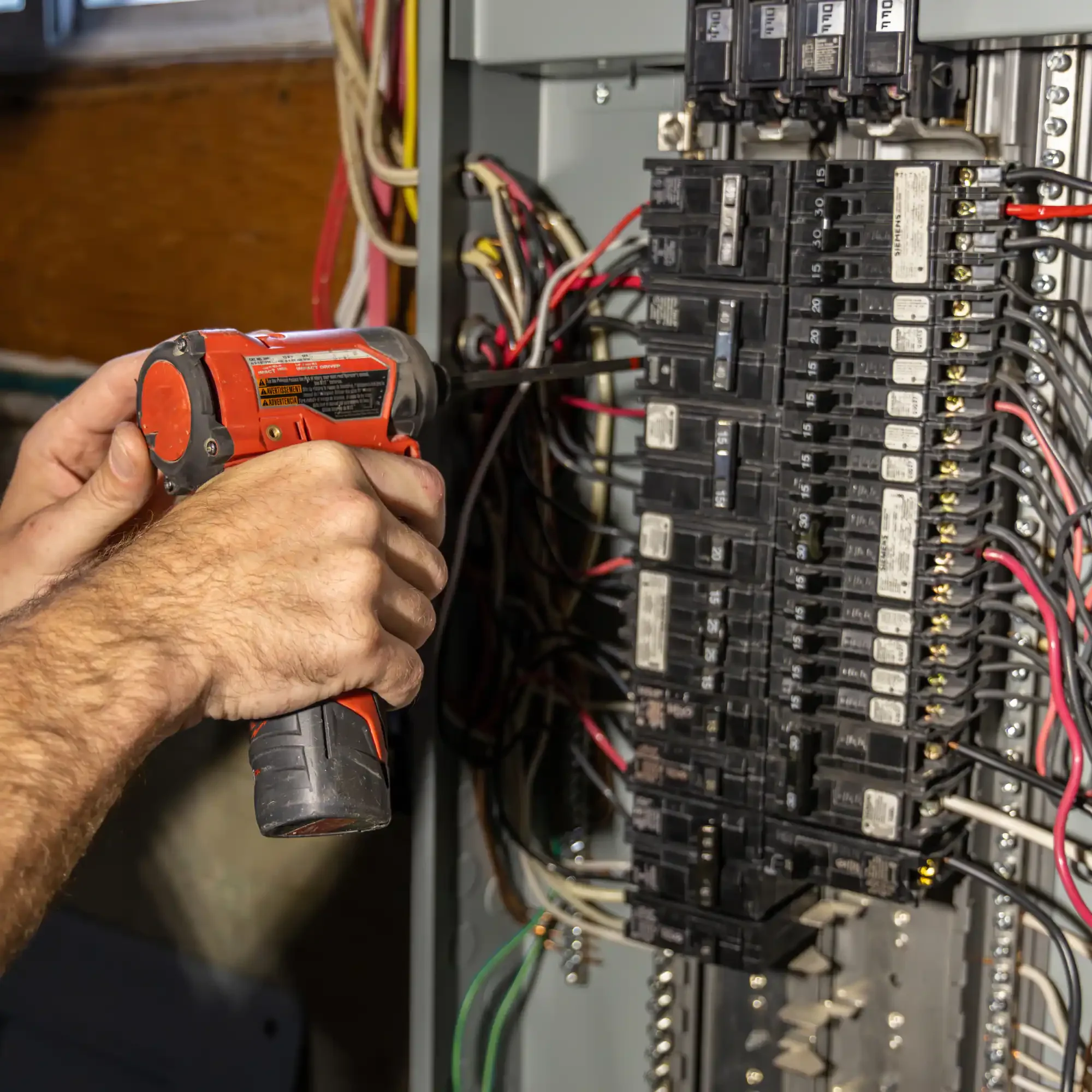 A person is using a power drill to work on an electrical breaker box with multiple circuit breakers.