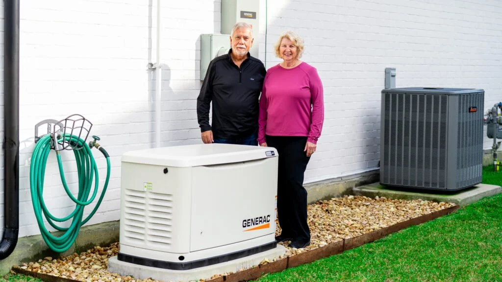 Older man and woman standing outside next to a Generac generator, green garden hose, and an air conditioning unit.