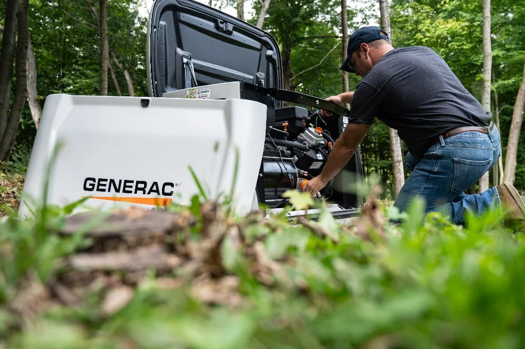 A man wearing a black t-shirt, blue jeans, and sunglasses kneeling in a forested area, inspecting or working on a large portable generator labeled 'Generac' with its open access panel visible.