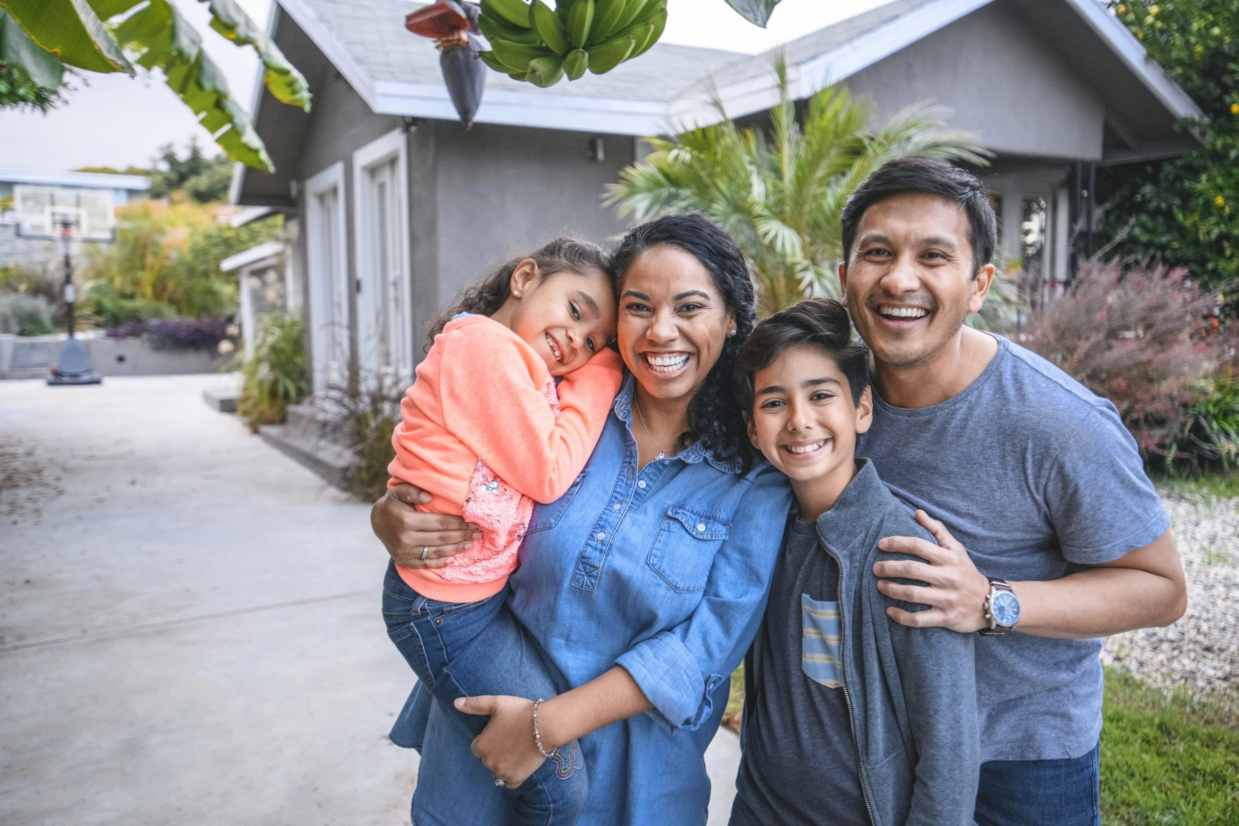 A happy family of four smiling and hugging outside in a driveway with a house and garden in the background.