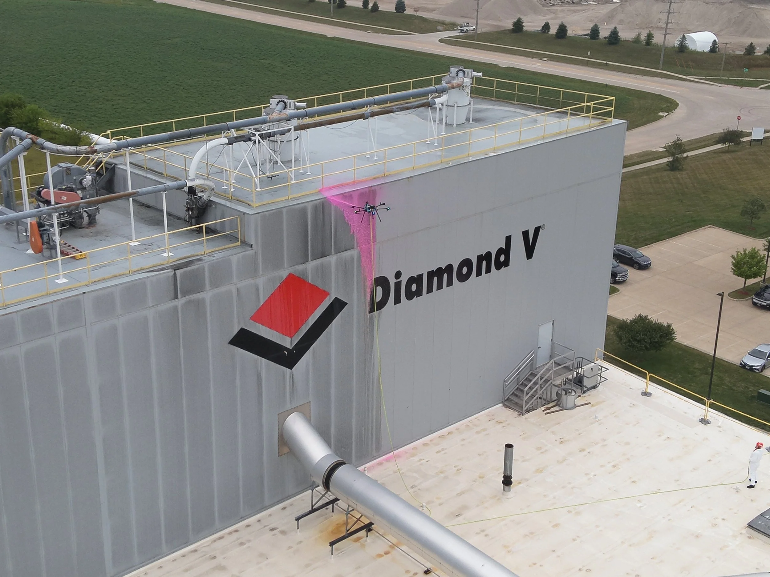 An industrial building with a large sign that reads 'Diamond V'. A drone is spraying a pink substance on the building's wall, with a person in protective gear visible on the ground nearby.