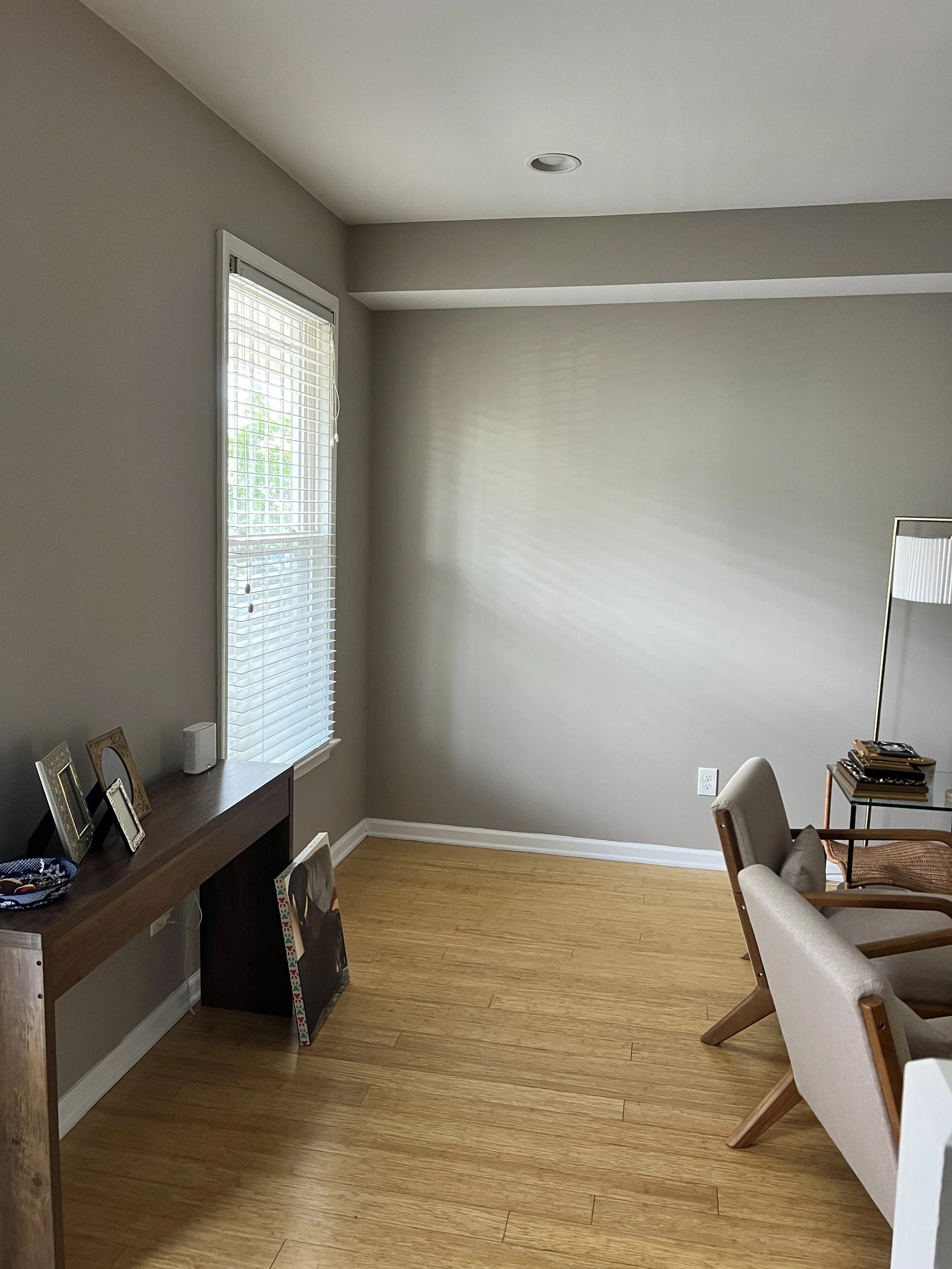 Interior room with hardwood floor, light-colored walls, a window with blinds, a wooden console table, framed pictures, a vase, and a sitting chair with a stack of magazines on a side table.