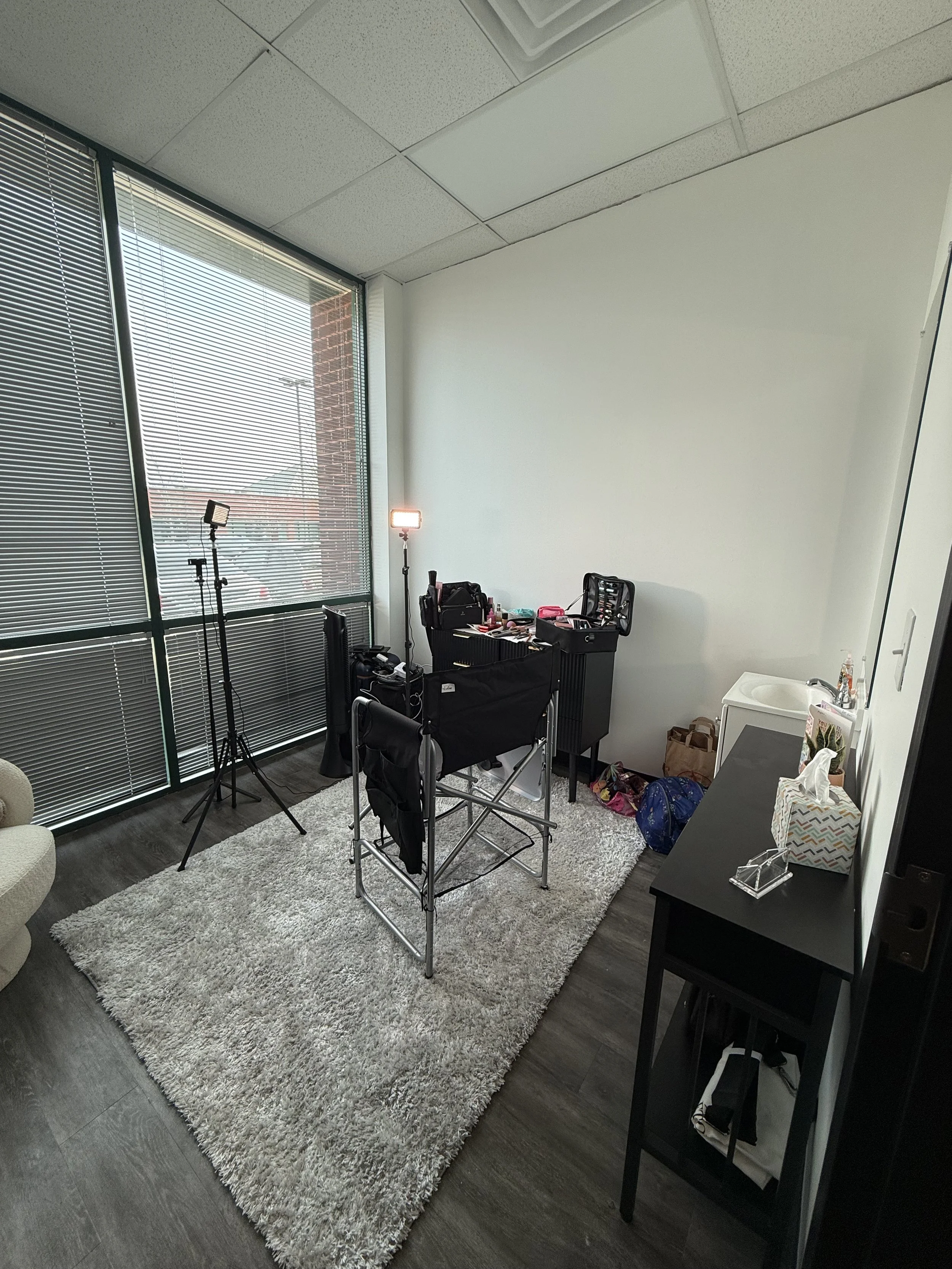 A room with a large window with blinds, a beige sofa armchair, a white fluffy rug, a black desk and a black dresser with tissue box, a mirror, and miscellaneous items, and studio lighting equipment.