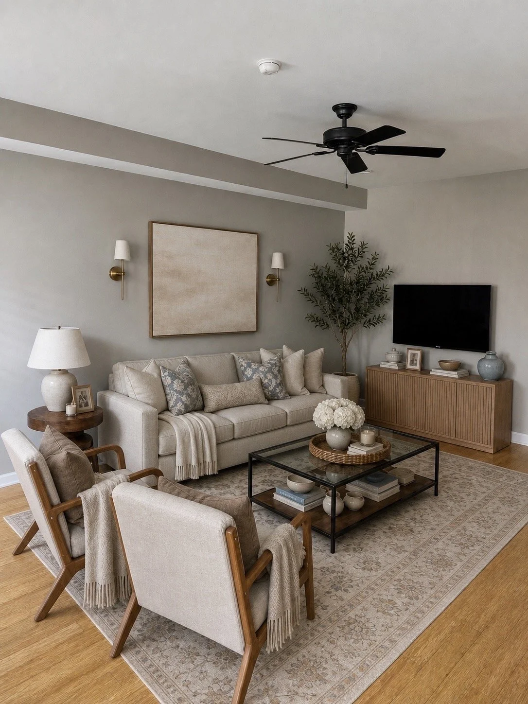 A living room with a beige sofa, armchairs, a glass coffee table, a large wall-mounted TV, and a wooden console against the wall. Decor includes a large framed art piece, wall sconces, table lamps, a potted plant, and decorative items, with a neutral