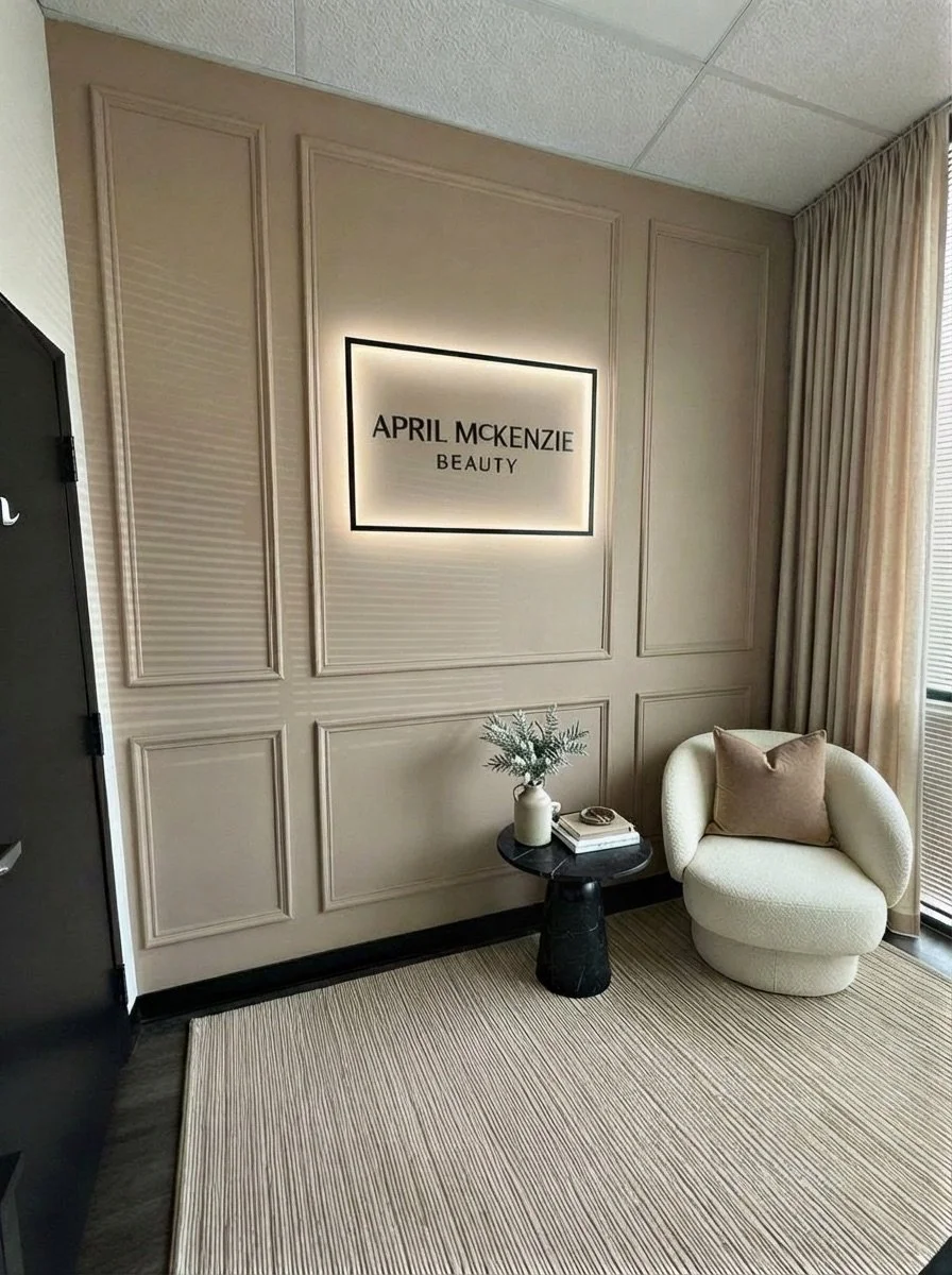 Interior of a beauty salon lounge with a beige wall, a backlit sign that reads 'April McKenzie Beauty,' a small black table with a vase and greenery, and a modern cream-colored chair with a brown pillow near floor-to-ceiling curtains.