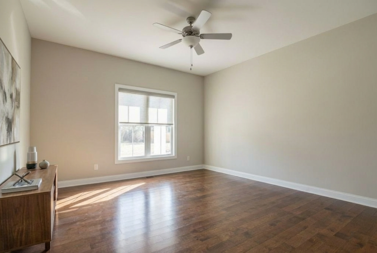 Empty living room with hardwood floors, light-colored walls, a ceiling fan, a window, and a wooden side table.
