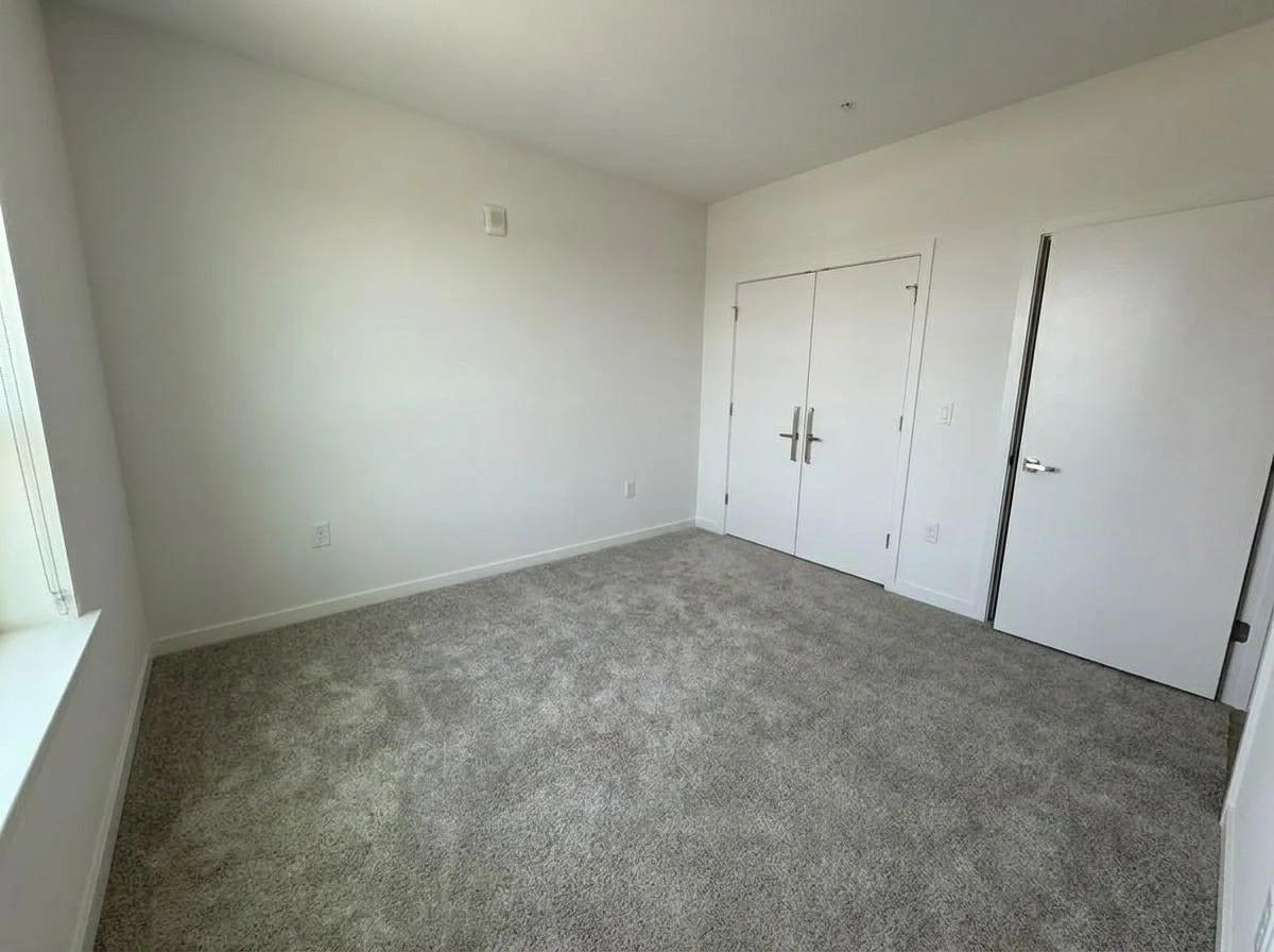 Empty room with white walls, gray carpet, a window with blinds, and double doors, in a residential property.