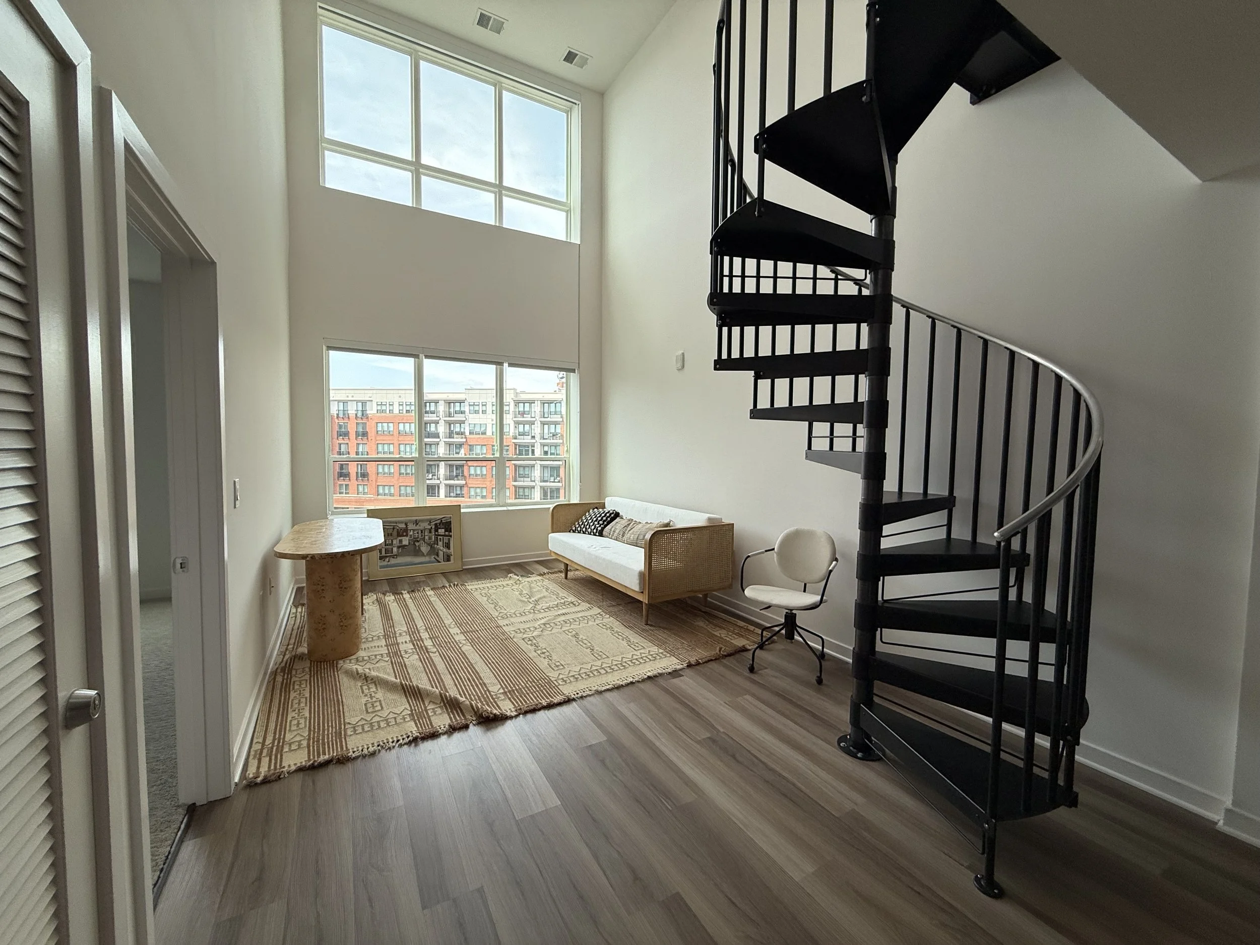 Living room with large windows, a beige rug, a small round table, a cream-colored sofa, and a black spiral staircase.