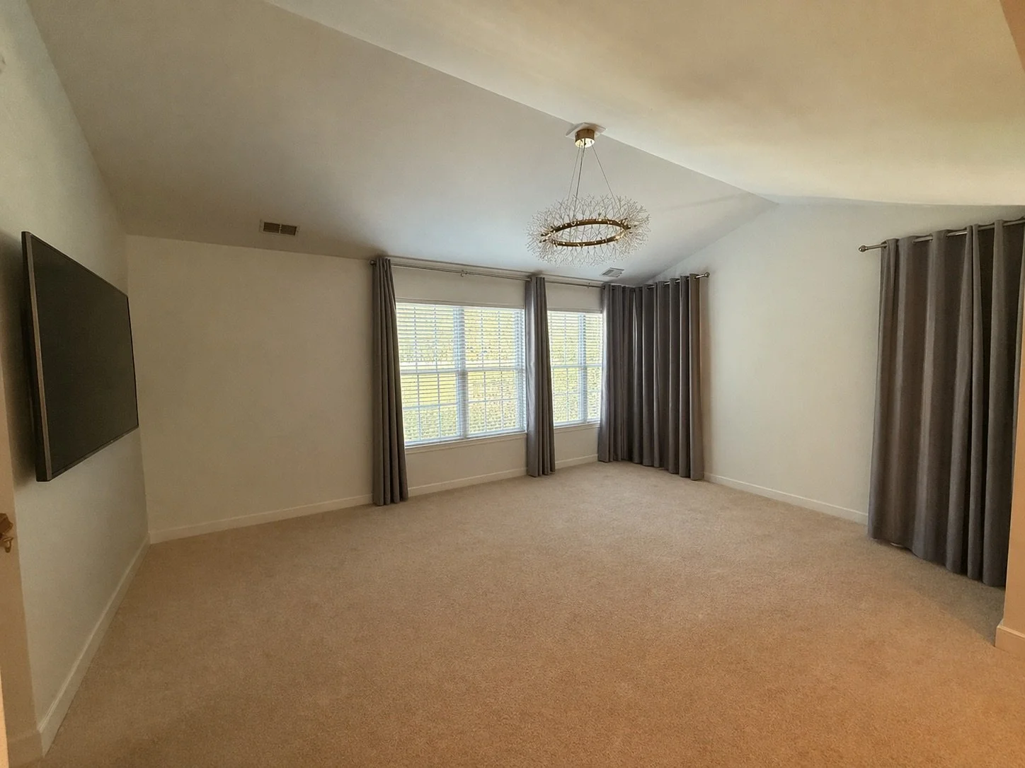 Empty living room with beige carpet, white walls, three large windows with blinds, gray curtains, a modern chandelier, and a wall-mounted flat-screen TV.