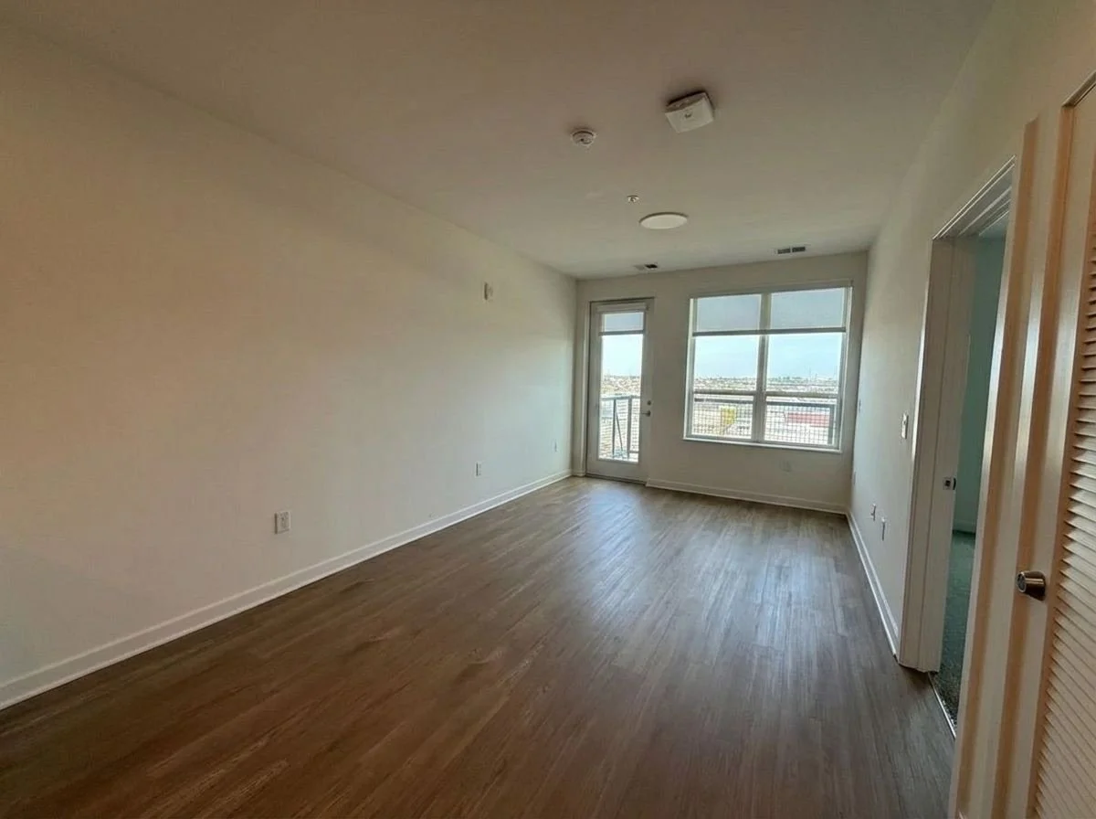 Empty apartment living room with hardwood floors, large windows, a door leading to a balcony, and a partially open closet door.