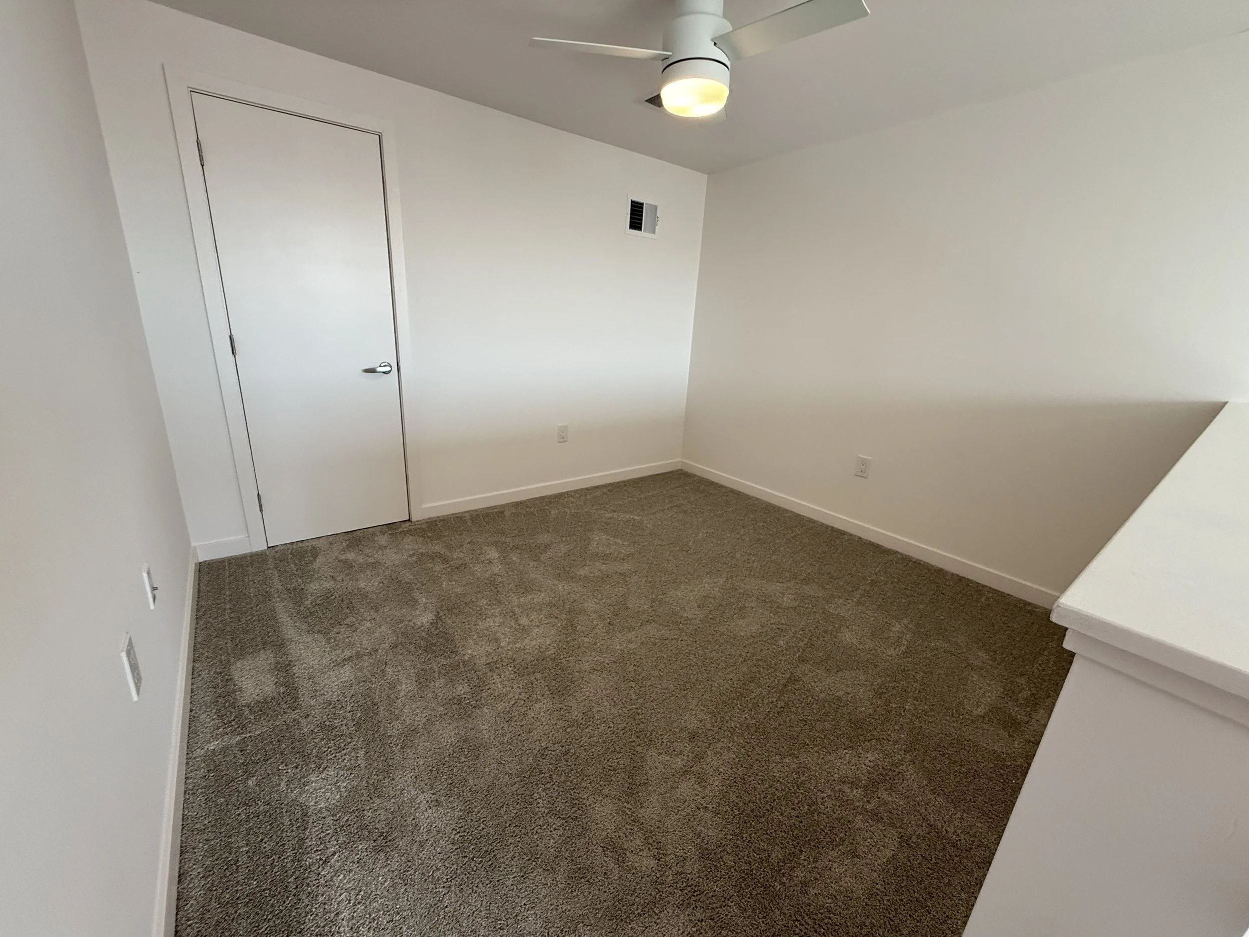 Empty room with white walls, a closed white door, ceiling fan with light, vents, and carpeted floor.