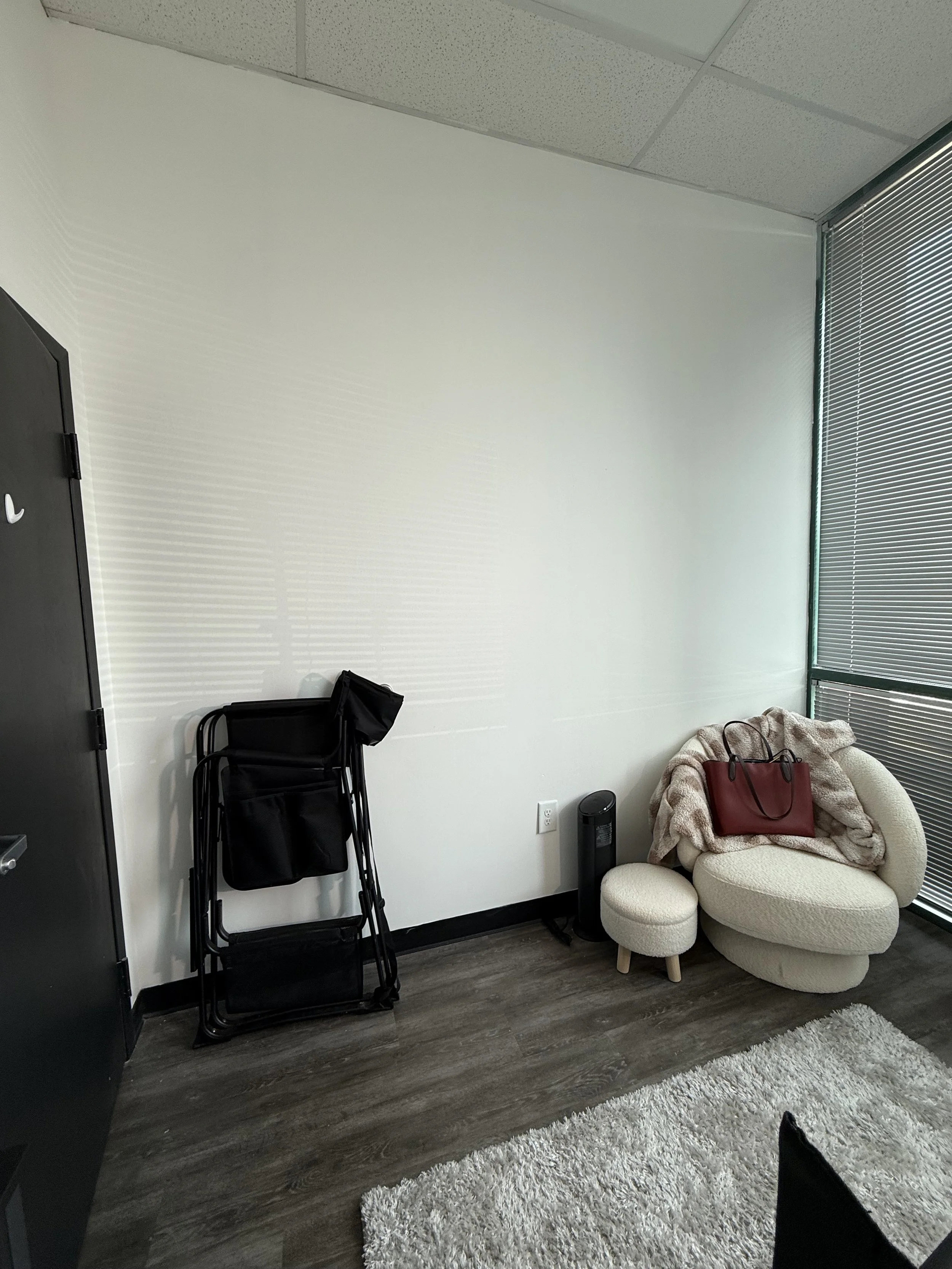 A corner of a room with a black door, a folded black chair, a beige sofa with a red bag and a blanket on it, a small round beige ottoman, a black air purifier, a white shaggy rug, and window blinds