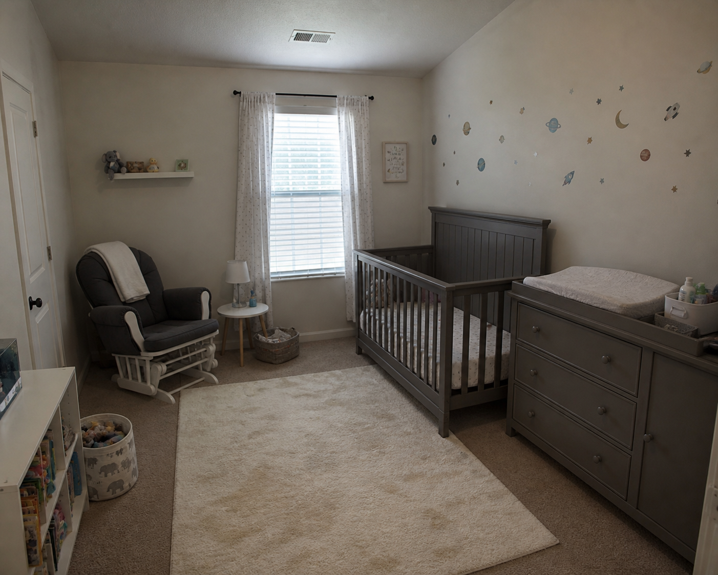 A nursery with a gray crib, a changing table, a gray rocking chair with a white pillow, a small white side table with a lamp, an area rug, a window with white curtains, and wall decals of planets, moons, stars, and rockets.