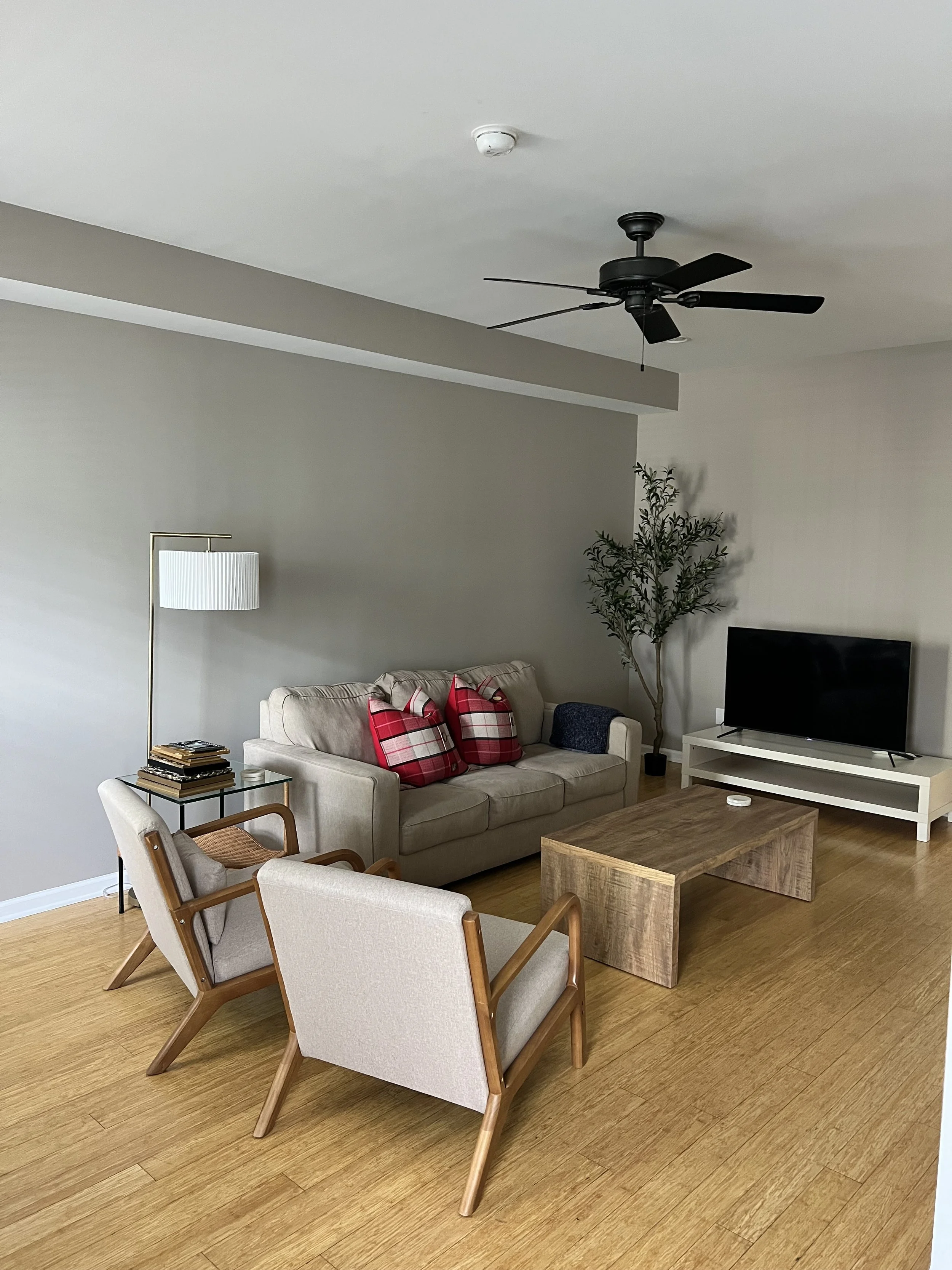 Living room with beige sofa, two armchairs, a black TV on a white stand, a wooden coffee table, a tall floor lamp with a white shade, a plant in the corner, and hardwood flooring.