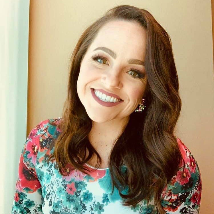 A woman with long, wavy brown hair and light makeup smiling, wearing a floral top with pink, blue, and black colors and large earrings.