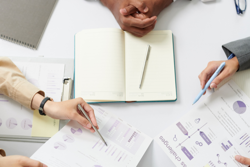 People working together at a desk with documents, charts, notebooks, and pens.