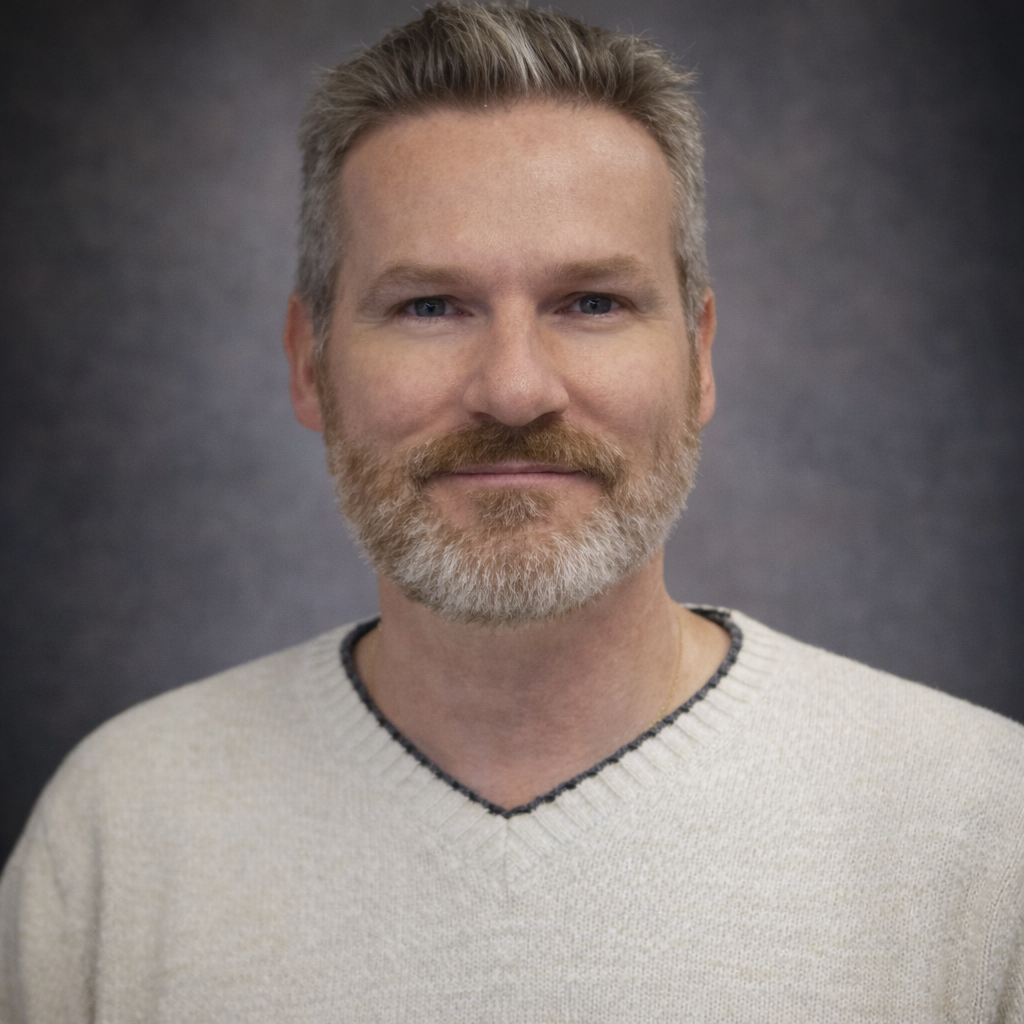 Instructor Joe Waters. A middle-aged man with a light beard and short gray hair smiling slightly, wearing a cream-colored sweater with a dark trim around the neckline, against a dark, neutral background.