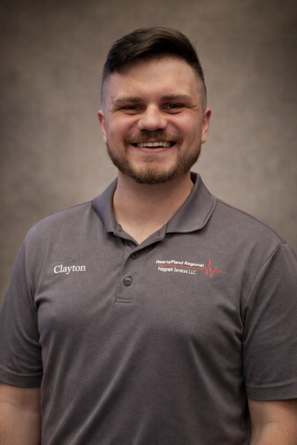 Lab Assistant Clay McNabb. A smiling man with short brown hair and a beard wearing a dark gray polo shirt. The background is a plain textured wall.