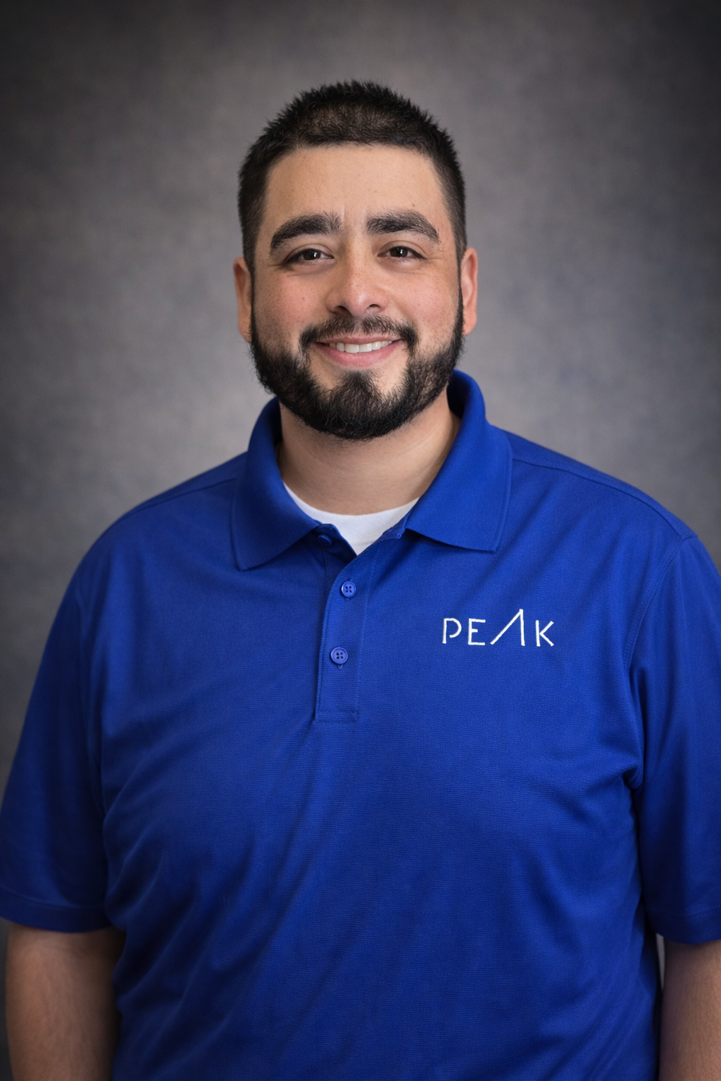 Lab Assistant Juan Navarro. A young man with a beard and short dark hair smiling, wearing a blue polo shirt with the word PEAK embroidered on it, standing against a gray background.