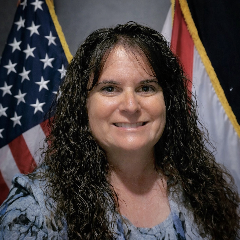 Director Sandy Dunn who is a woman with curly brown hair, standing in front of American and state flags.