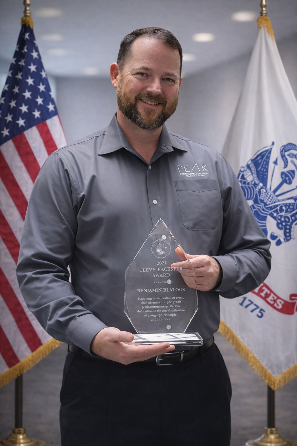 Consulting Partner Ben Blalock. A man with a beard smiling, wearing a gray shirt, is holding a clear glass award with an inscription. Behind him are American and organizational flags.