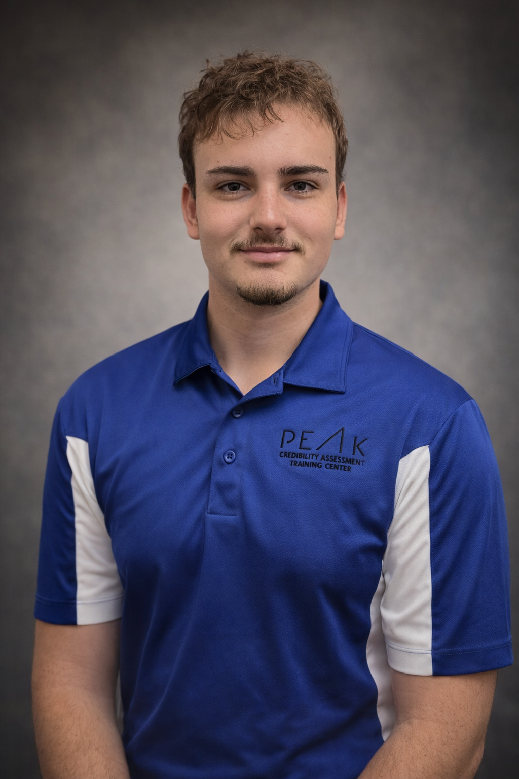 Lab Assistant Niko Scherer. A young man with light brown hair and a goatee, wearing a blue polo shirt with white accents on the sleeves, standing against a gray background.