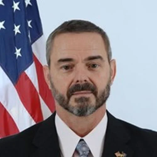 Senior Instructor Jim Orr. Portrait of a man with a beard and short hair, wearing a suit and tie, standing in front of a United States flag.