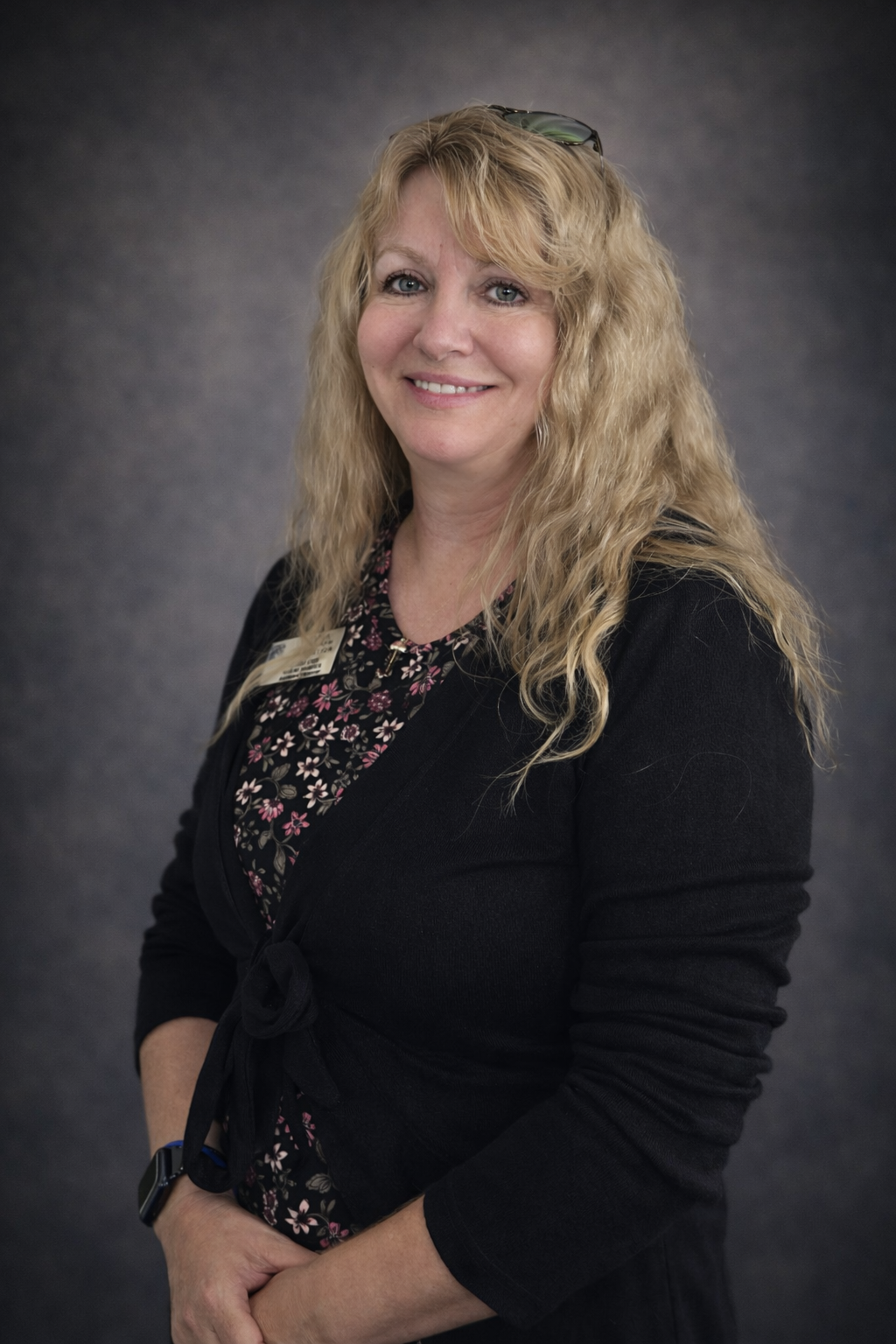 Lab Assistant Trish Orr. A woman with long, curly blonde hair smiling, wearing a black blazer and floral blouse, with glasses on her head and a smartwatch on her wrist, standing against a dark gray background.